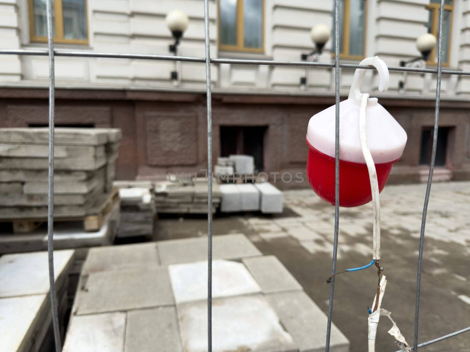 Close up of red construction warning light hanging on temporary metal fence with stacked paving tiles in background. The image reflects active urban renewal and safety measures