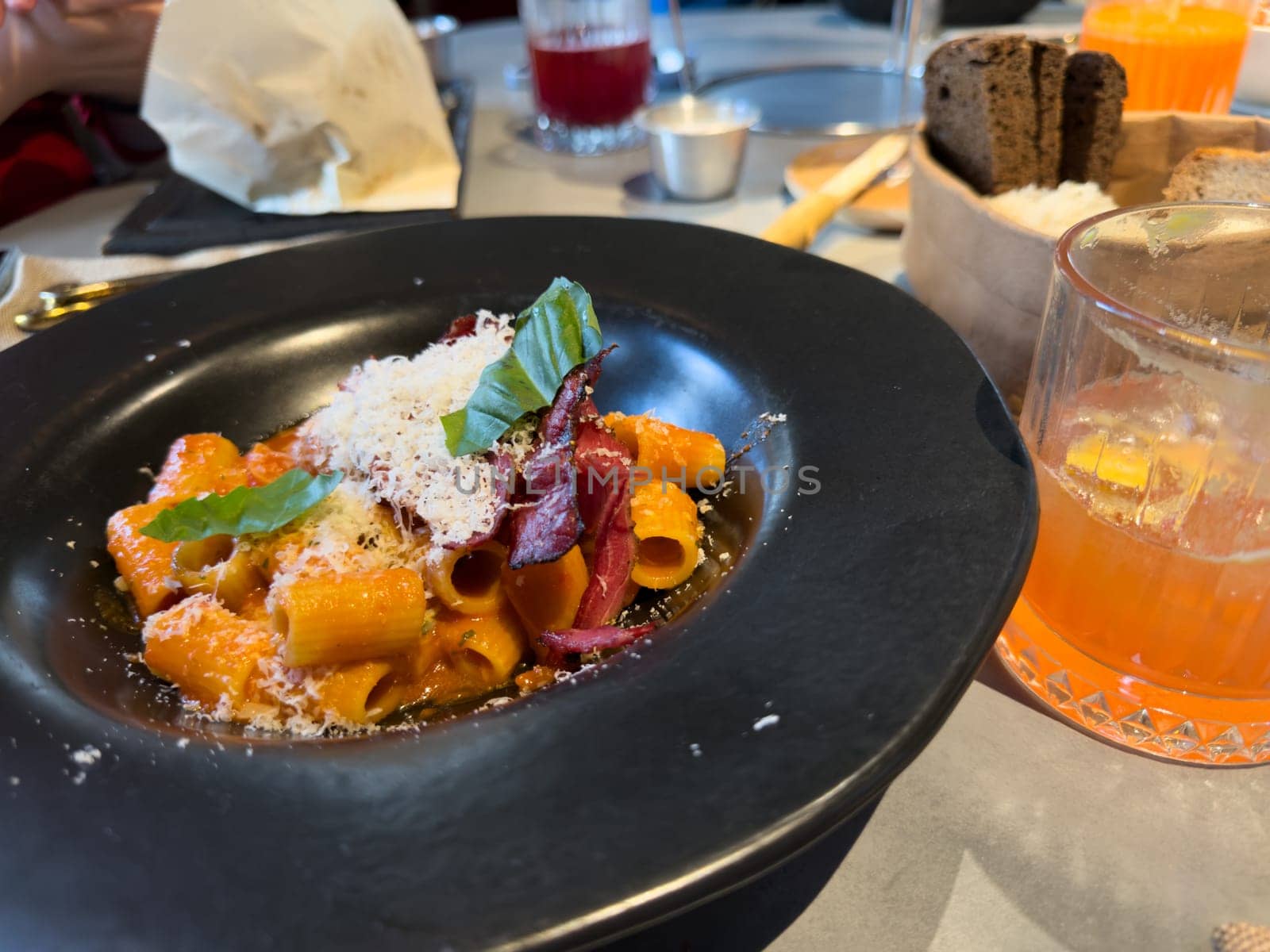 Plate of rigatoni pasta with tomato sauce, crispy bacon, grated cheese and fresh basil leaves. Served in modern restaurant setting with orange drink and sliced bread on the side