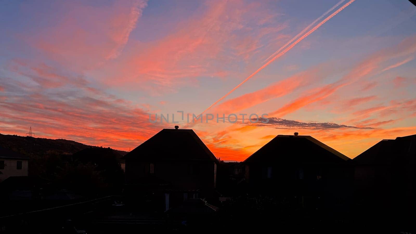 A stunning sunset illuminates the sky with shades of orange and pink over suburban rooftops. Vapor trails create accents across this serene urban landscape, offering a picturesque view.