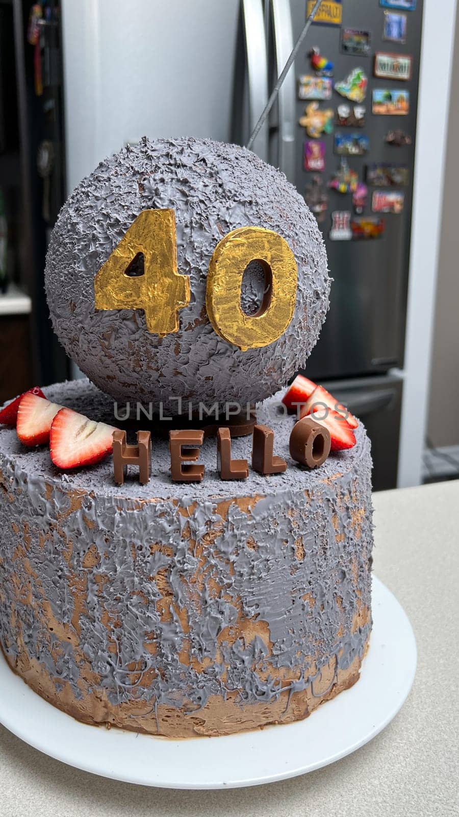 A stylish cake for a 40th birthday. It has grey textured frosting, golden numbers, chocolate letters that say HELLO, and fresh strawberries for a delightful finish. Perfect for the celebration.
