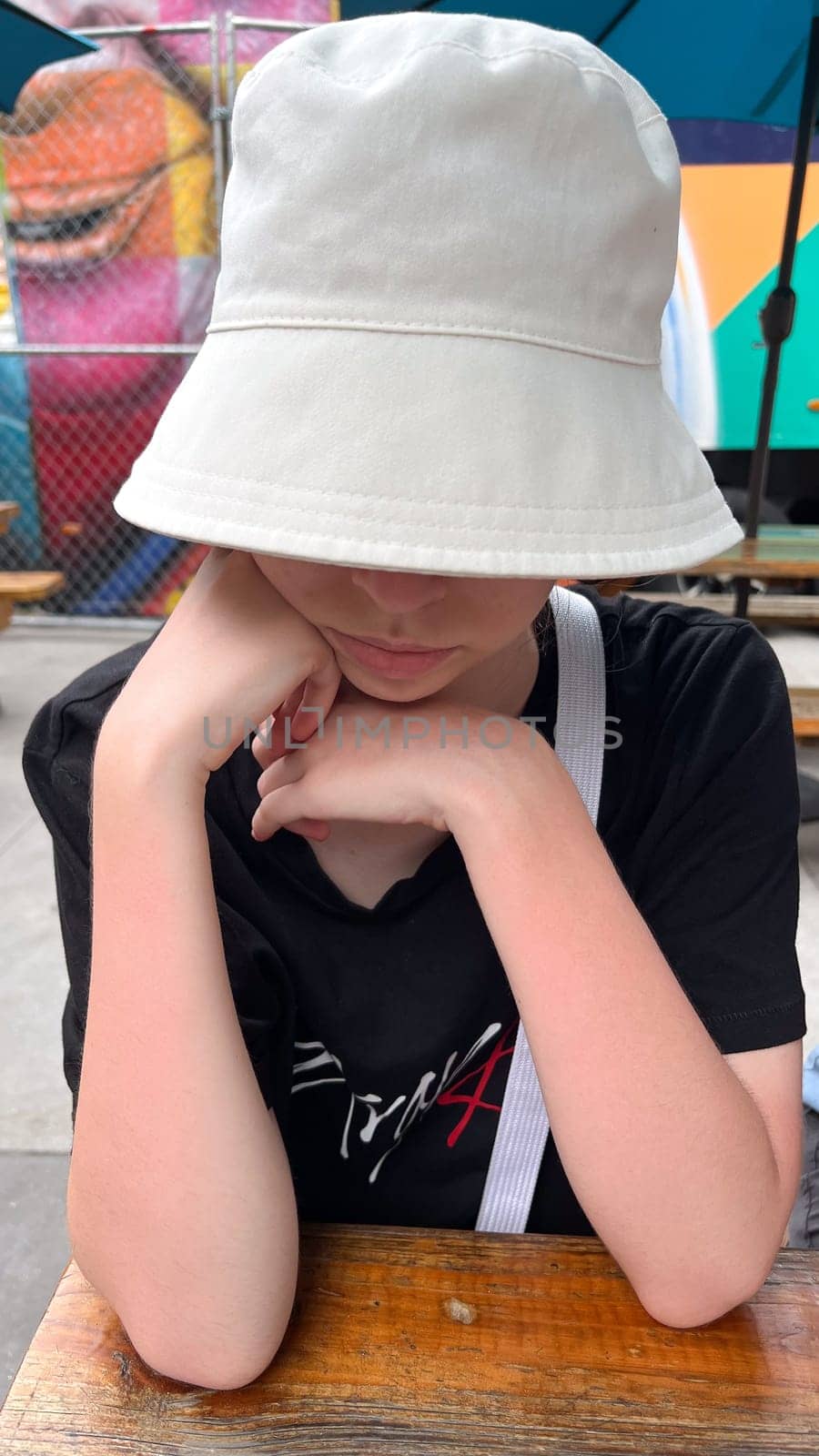 A young person in a casual bucket hat sits at a wooden table, appearing deep in thought. This image encapsulates themes of youth, introspection, and the simplicity of everyday life.