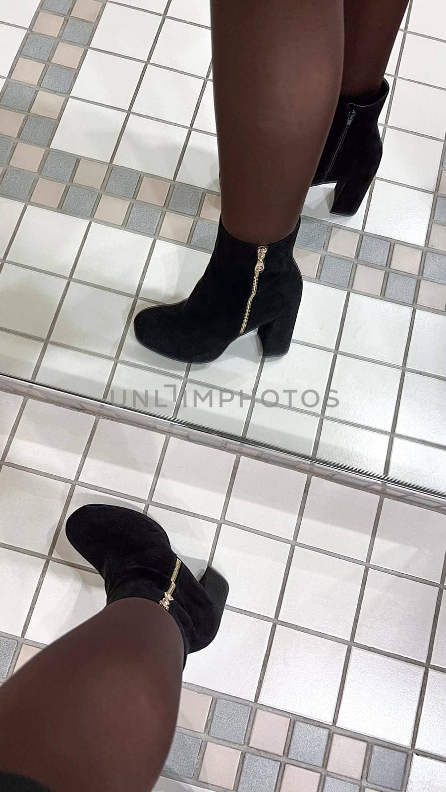 A chic black suede ankle boot with a gold zipper is reflected in a mirror on a tiled floor. This stylish boot is perfect for fashion designs, advertisements, and editorial content.