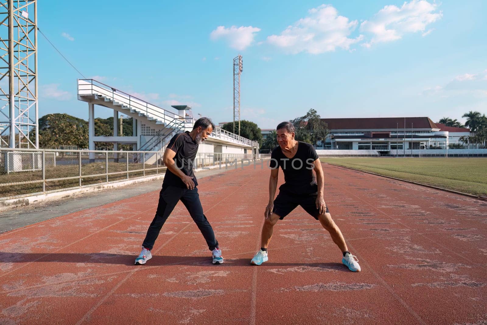 Two elderly men stretching together on the track, preparing for their workout and highlighting the importance of wellness and fitness in active aging.