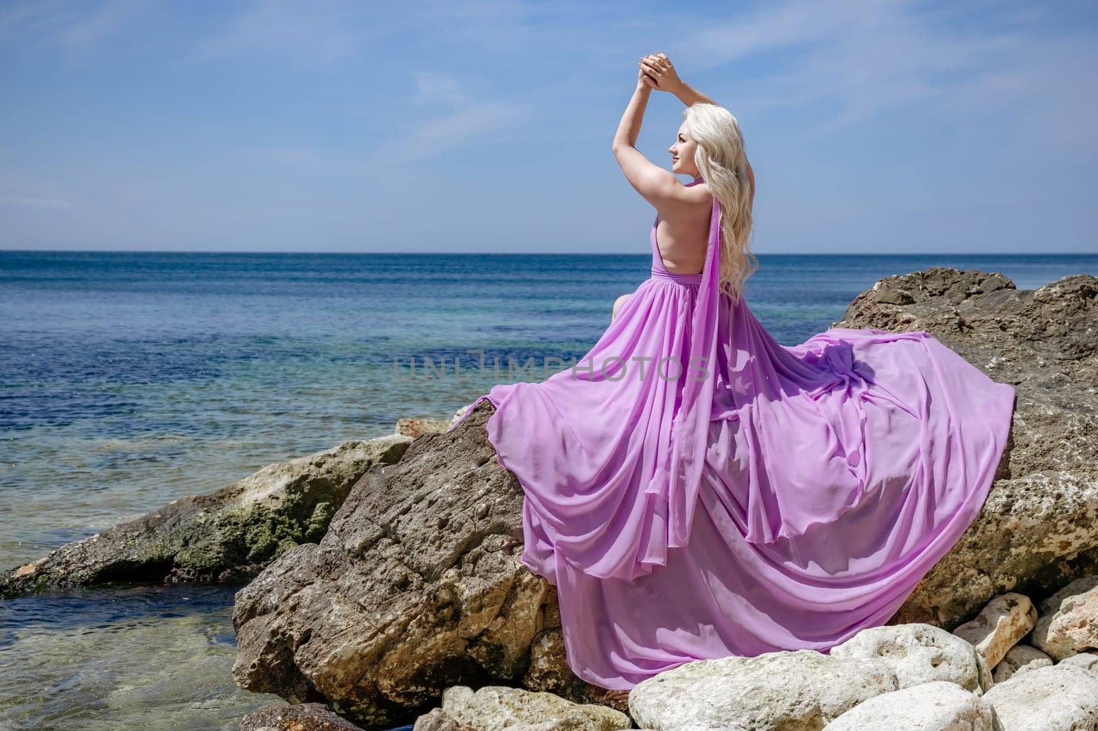 Blonde woman in flowing purple dress posing on rocky beach overlooking the sea. Back view.