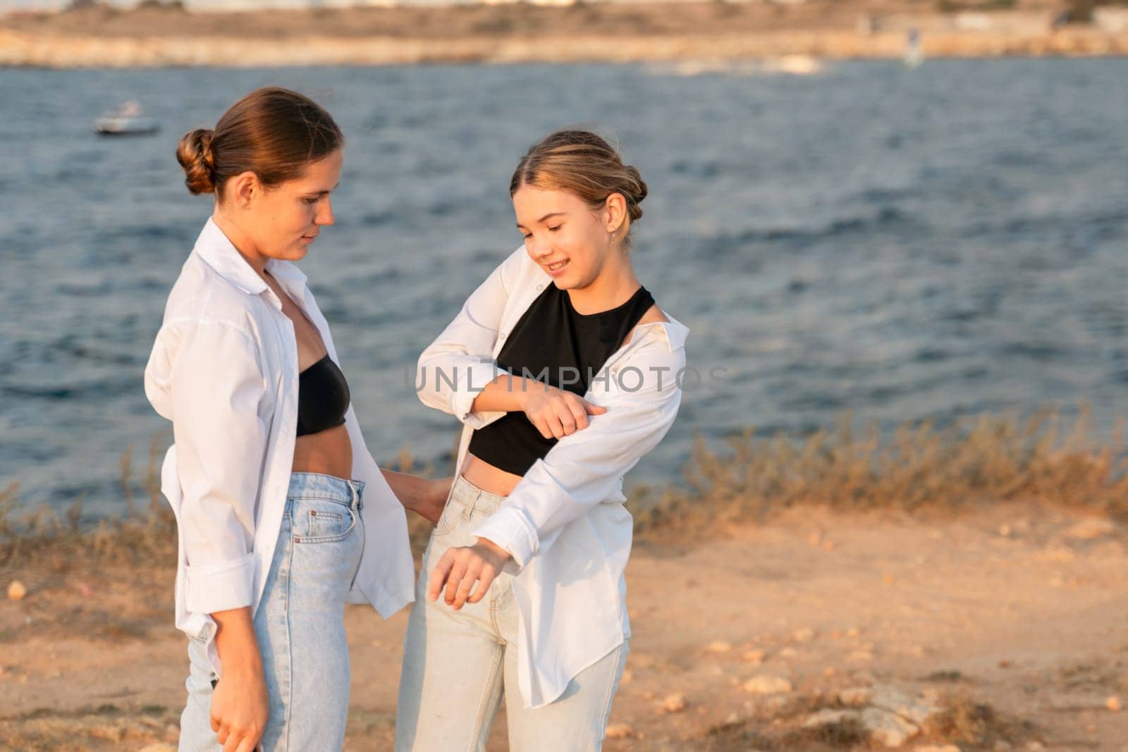 Two young women stand on a beach, one in a white shirt and jeans, the other in a black crop top and jeans. They are laughing and talking to each other.
