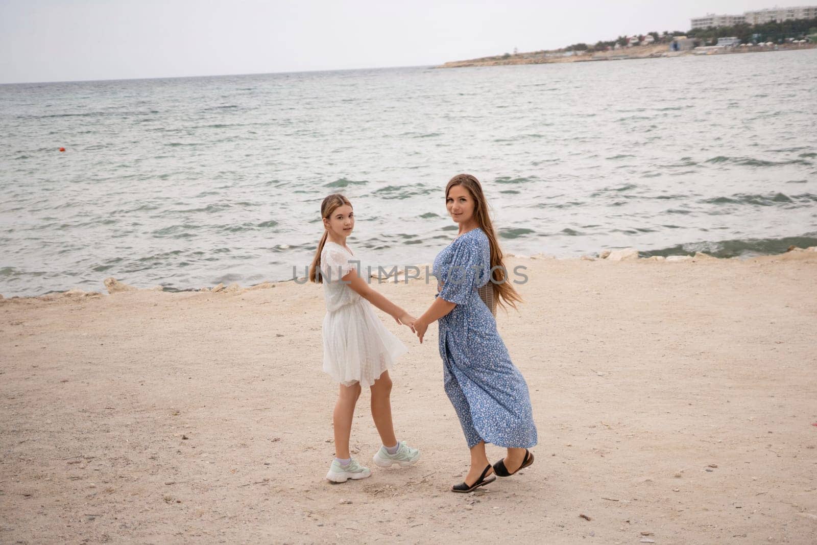 Two women, holding hands, stand on a sandy beach near a calm sea. One wears a white dress, the other a blue one. They are smiling.