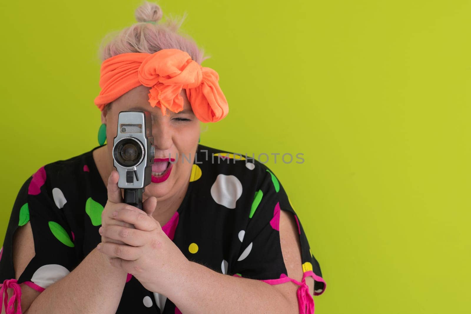 Playful and plus size woman with pink hair and orange headband aiming a vintage film camera, wearing a colorful polka dot dress against a vivid green background.