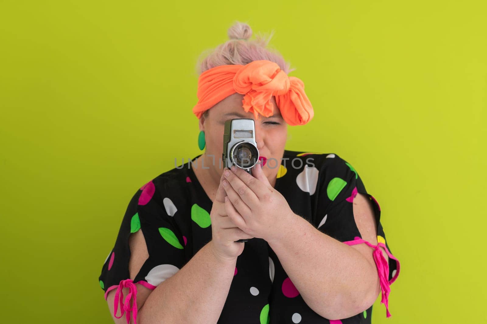 Playful and plus size woman with pink hair and orange headband aiming a vintage film camera, wearing a colorful polka dot dress against a vivid green background.