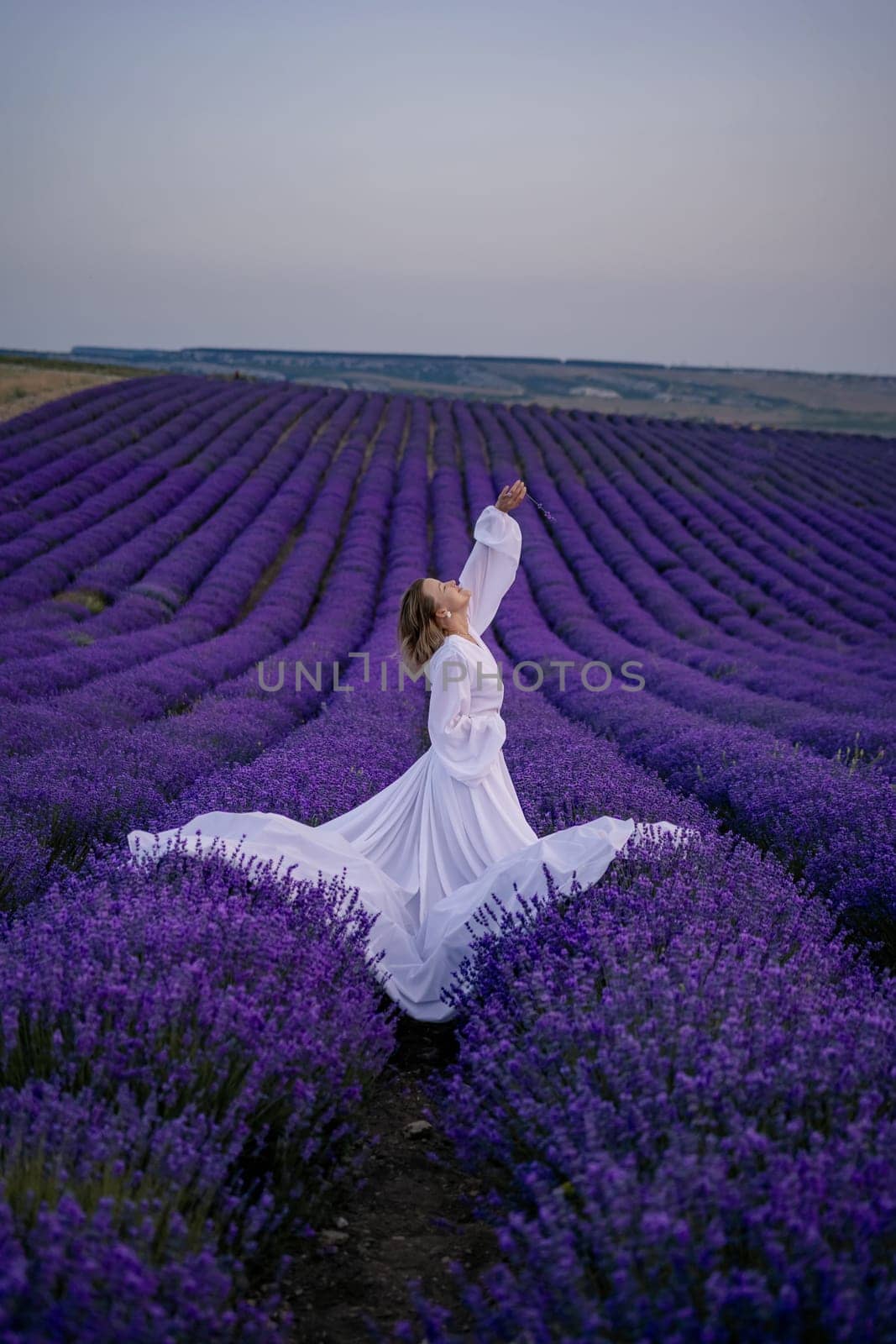 A woman in a white dress is standing in a field of purple flowers. She is reaching up with her arms outstretched, as if she is reaching for something