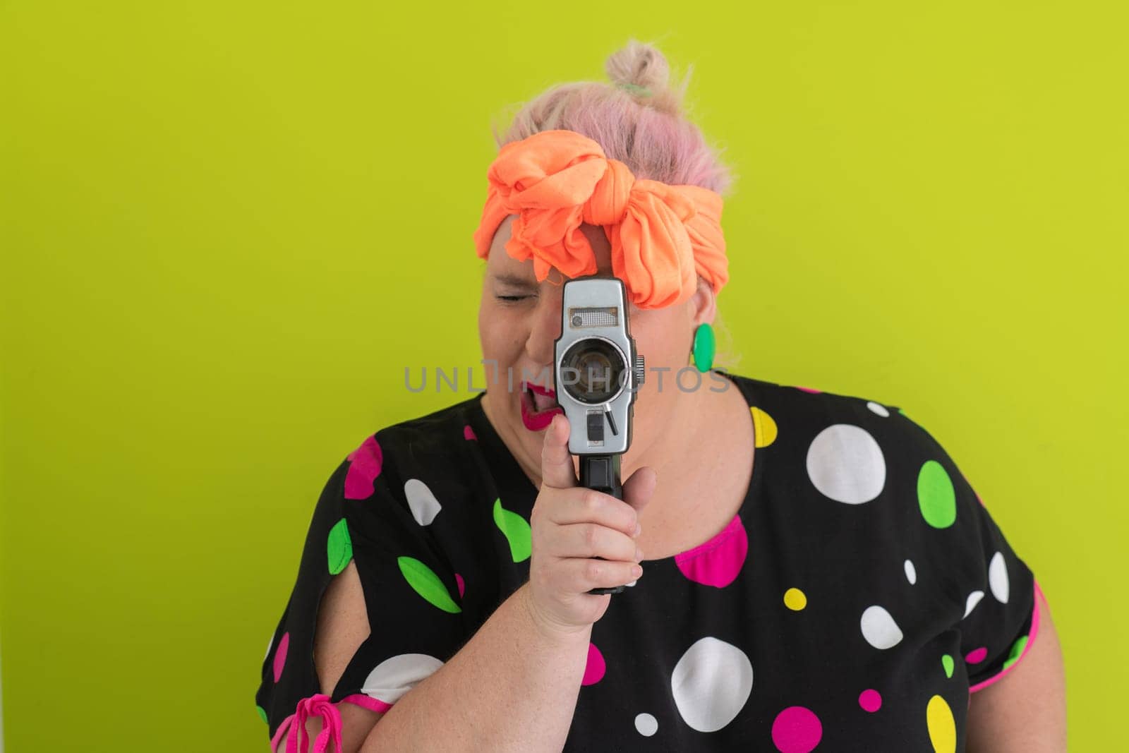 Playful and plus size woman with pink hair and orange headband aiming a vintage film camera, wearing a colorful polka dot dress against a vivid green background.