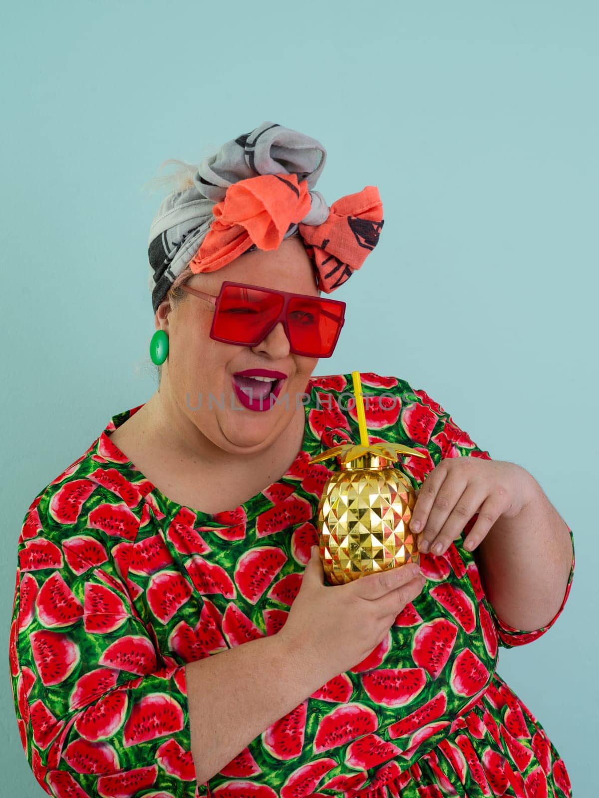 Eccentric plus size woman in a watermelon-patterned dress and colorful headwrap holding a golden pineapple-shaped cup with a straw, playfully glancing over her red sunglasses.