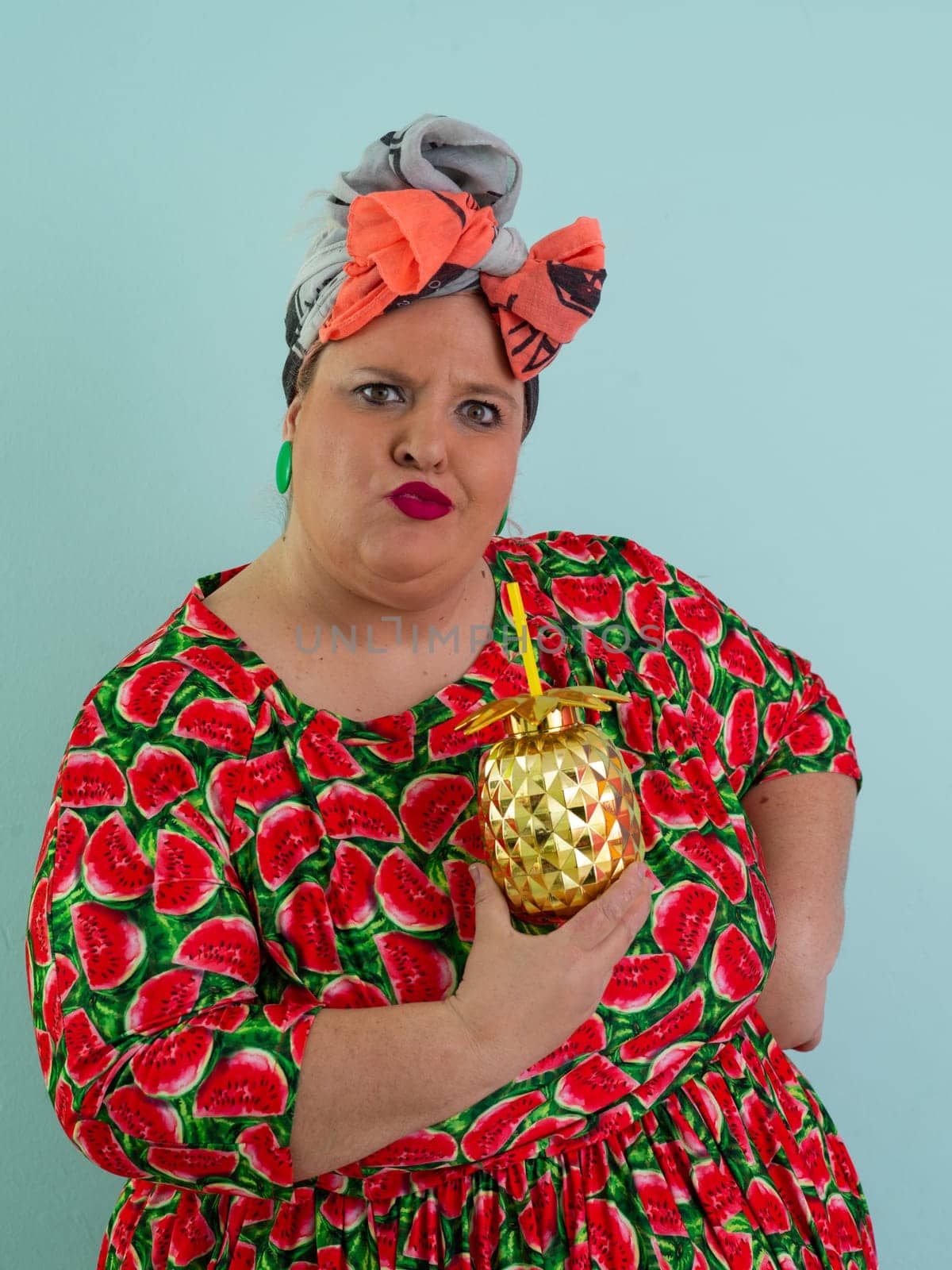 Eccentric plus size woman in a watermelon-patterned dress and colorful headwrap holding a golden pineapple-shaped cup with a straw, playfully glancing over her red sunglasses.