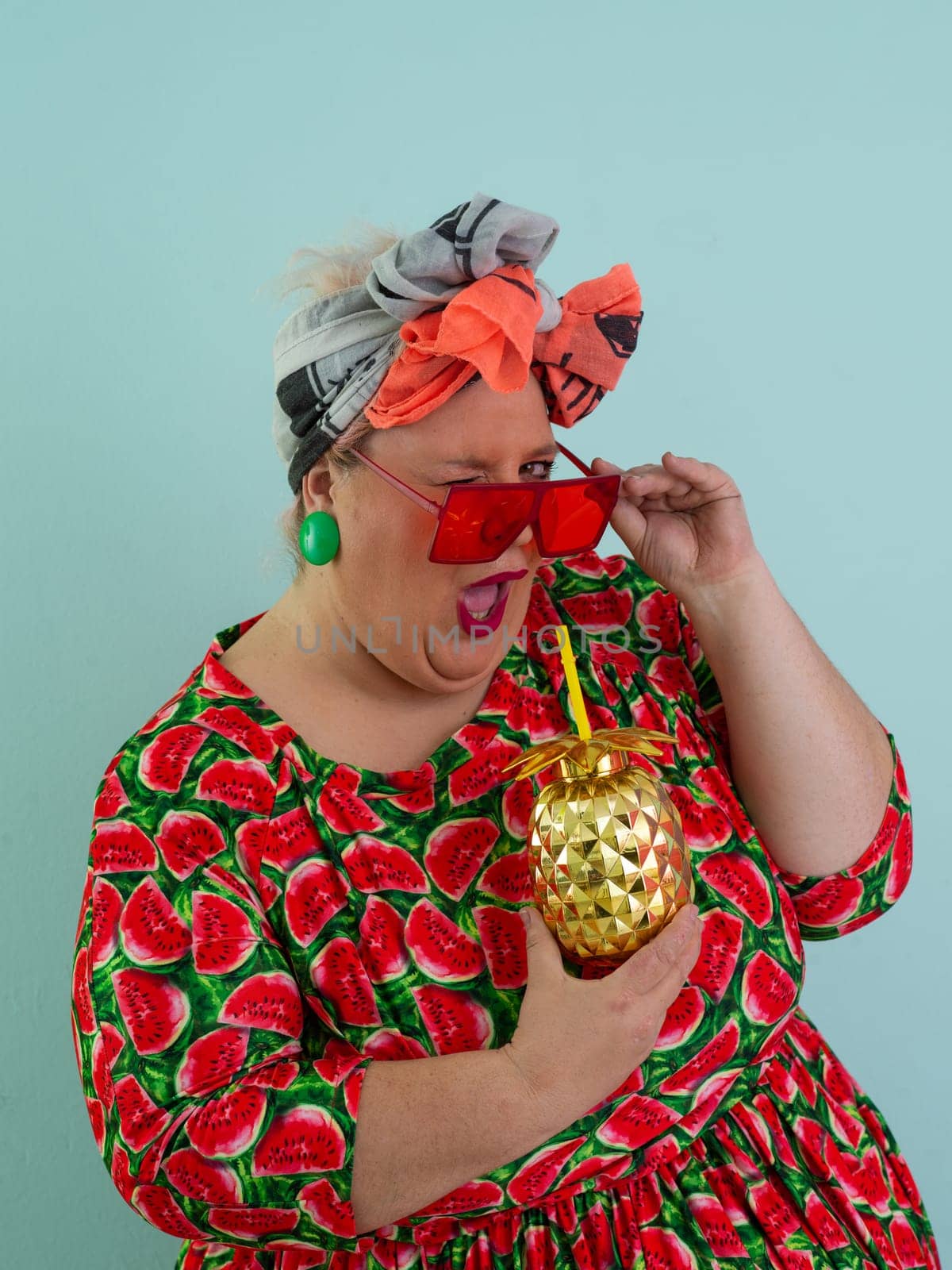 Eccentric plus size woman in a watermelon-patterned dress and colorful headwrap holding a golden pineapple-shaped cup with a straw, playfully glancing over her red sunglasses.