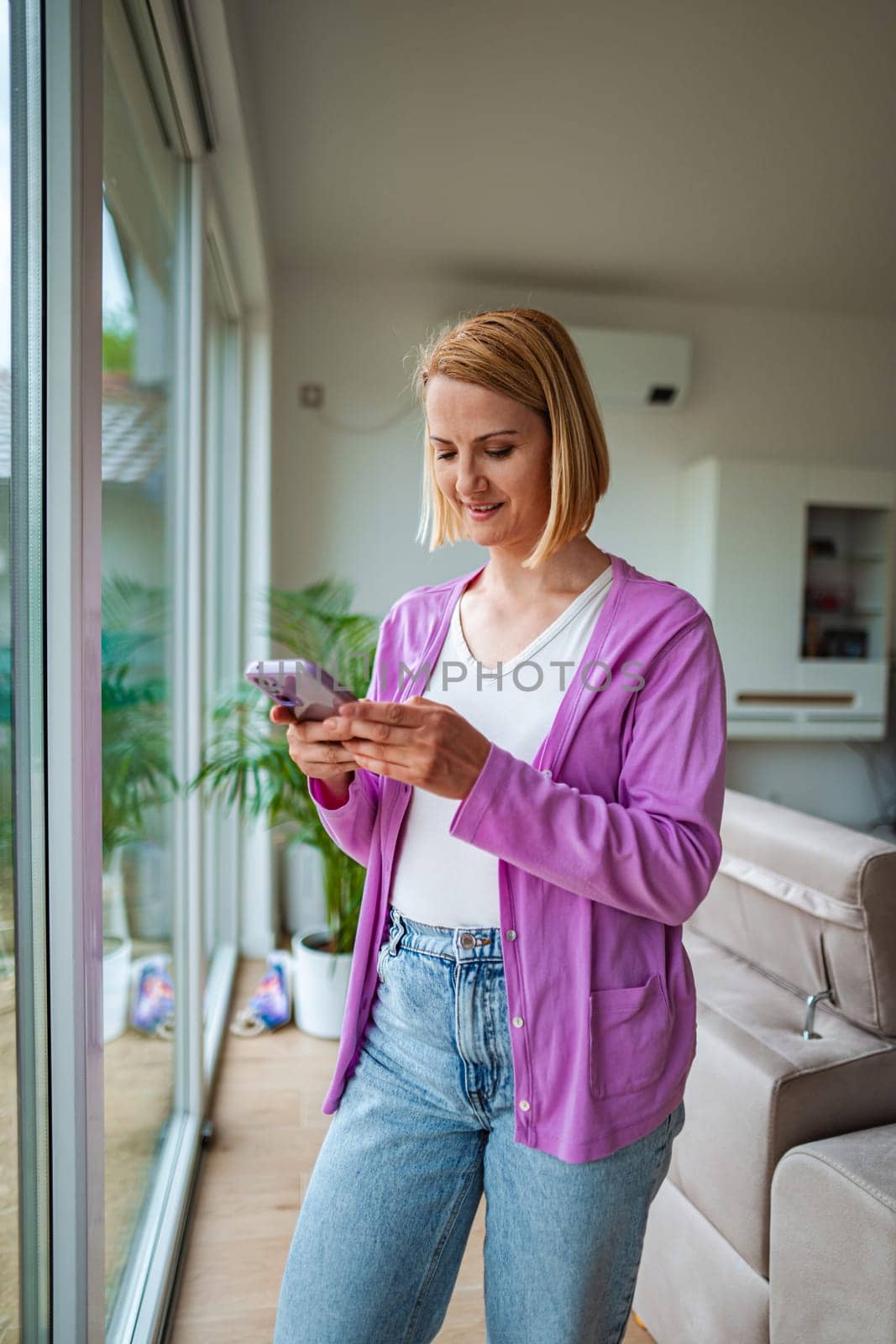 Young woman enjoys texting by a large window in her home.