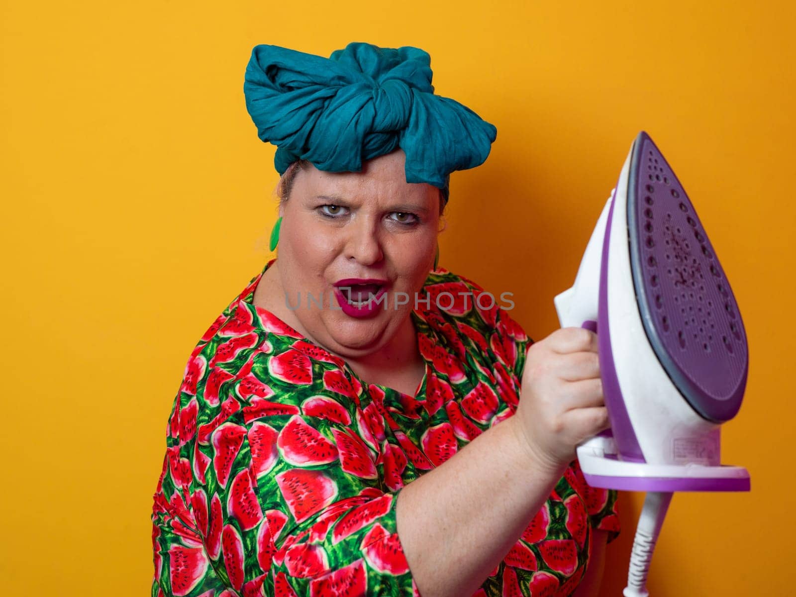 Plus size woman in a vibrant watermelon-patterned dress and teal headwrap, holding an electric iron and shouting with a humorous expression against a bright orange background. Conceptual and colorful portrait representing loudness, domestic chores, and quirky energy.