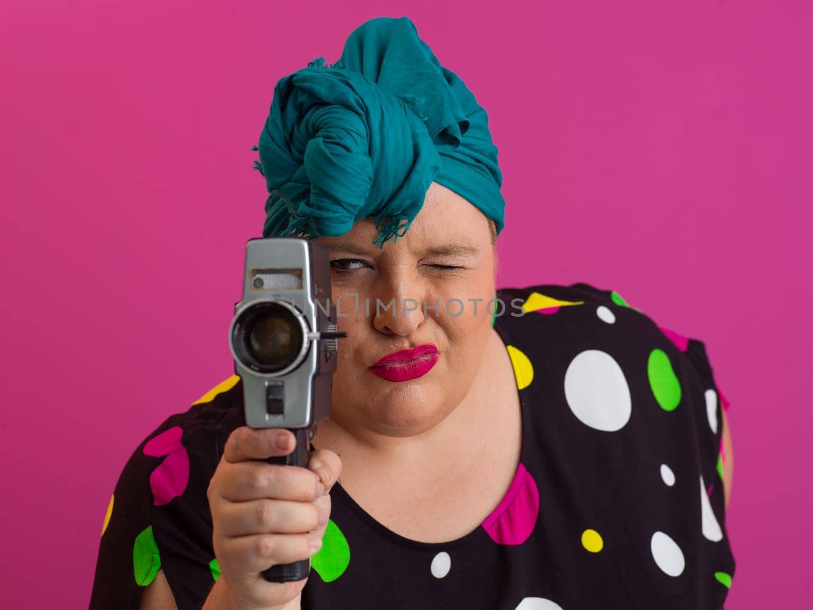 Playful and plus size woman with pink hair and orange headband aiming a vintage film camera, wearing a colorful polka dot dress against a pink background.