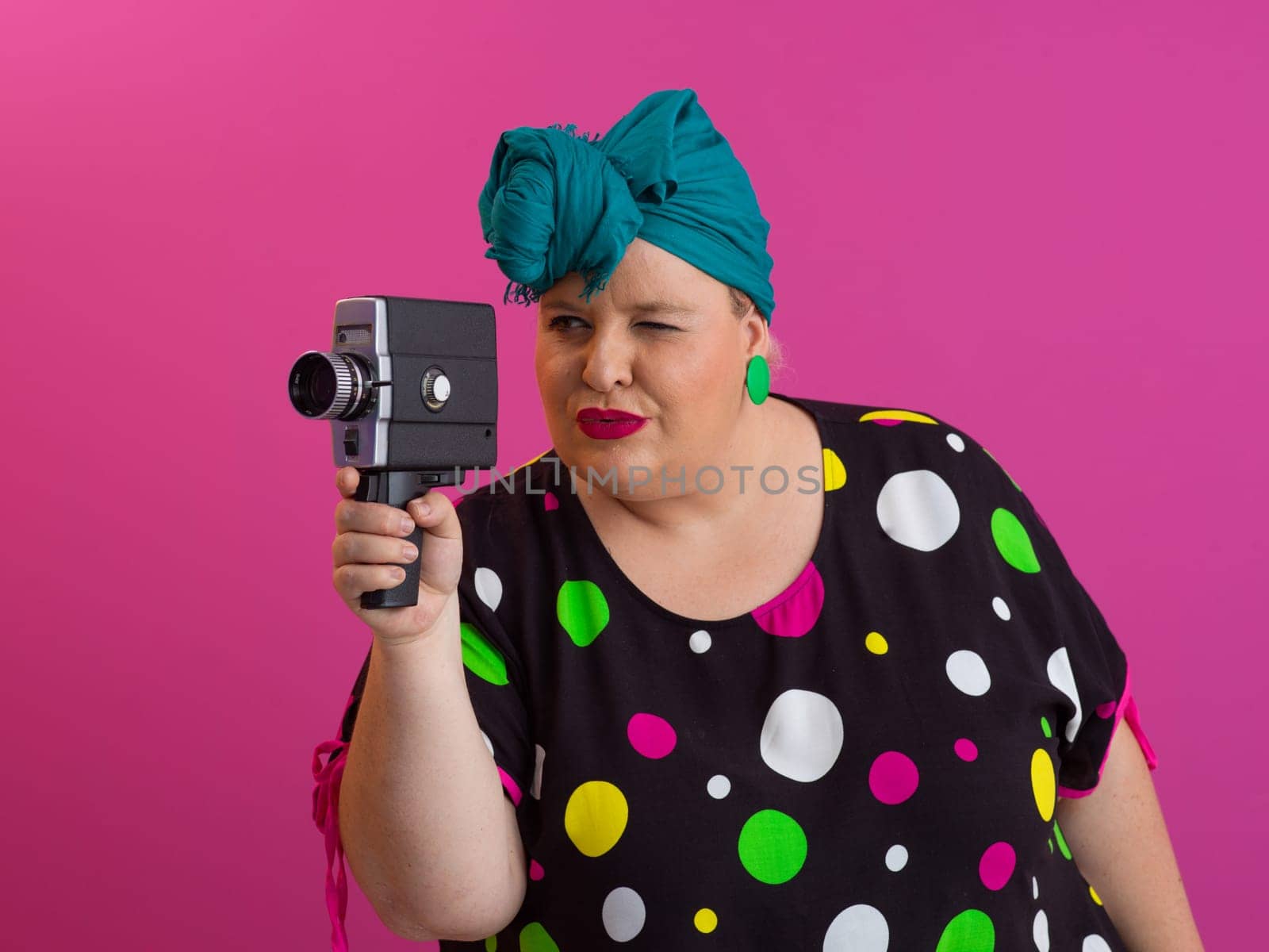 Playful and plus size woman with pink hair and orange headband aiming a vintage film camera, wearing a colorful polka dot dress against a pink background.
