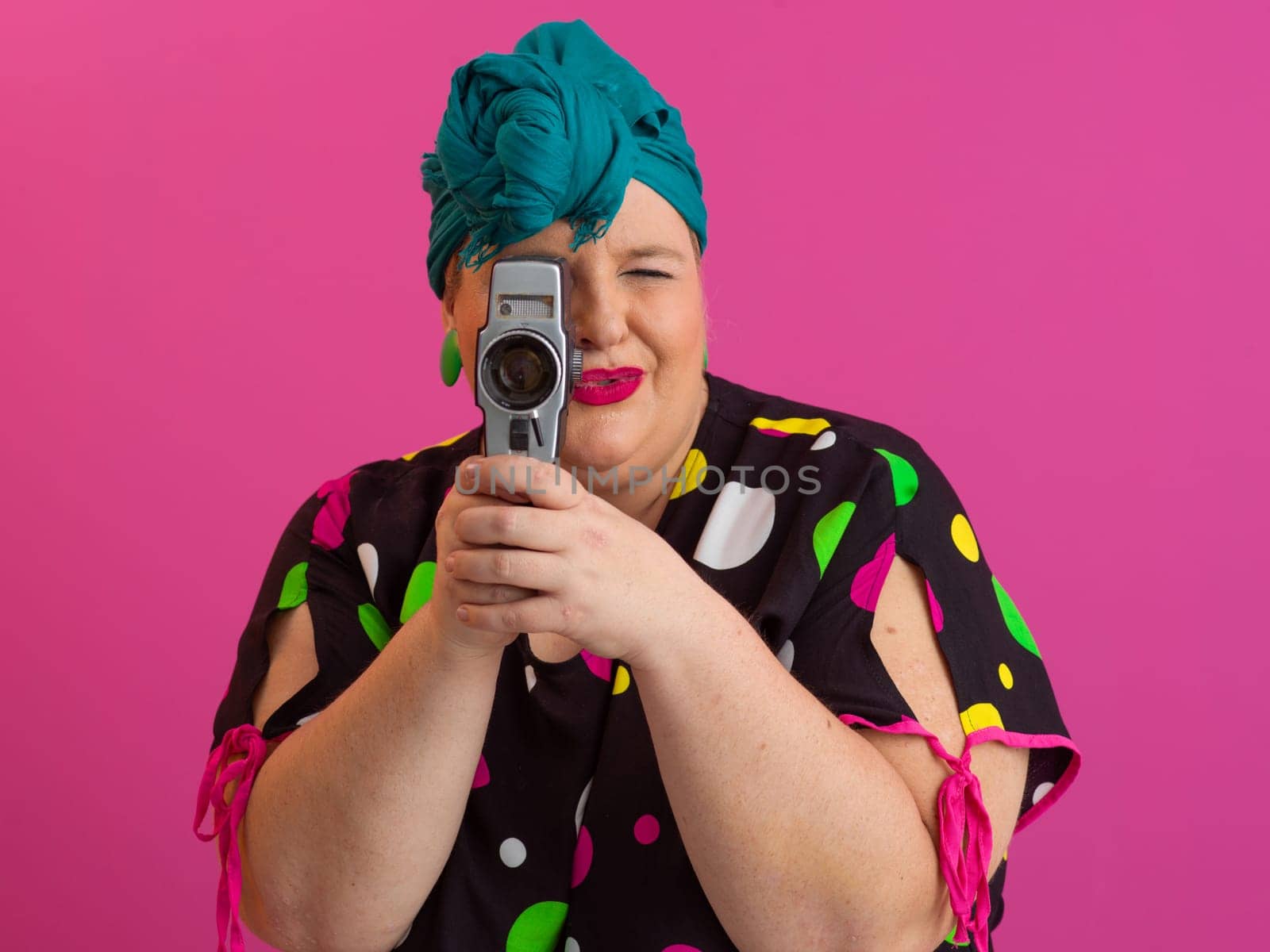 Playful and plus size woman with pink hair and orange headband aiming a vintage film camera, wearing a colorful polka dot dress against a pink background.