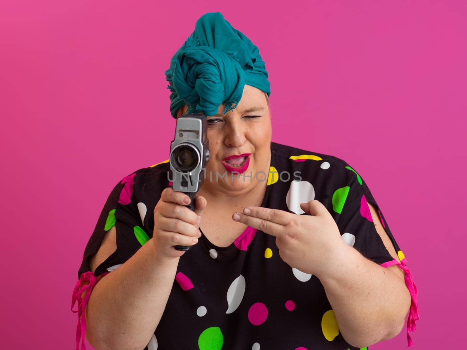 Playful and plus size woman with pink hair and orange headband aiming a vintage film camera, wearing a colorful polka dot dress against a pink background.