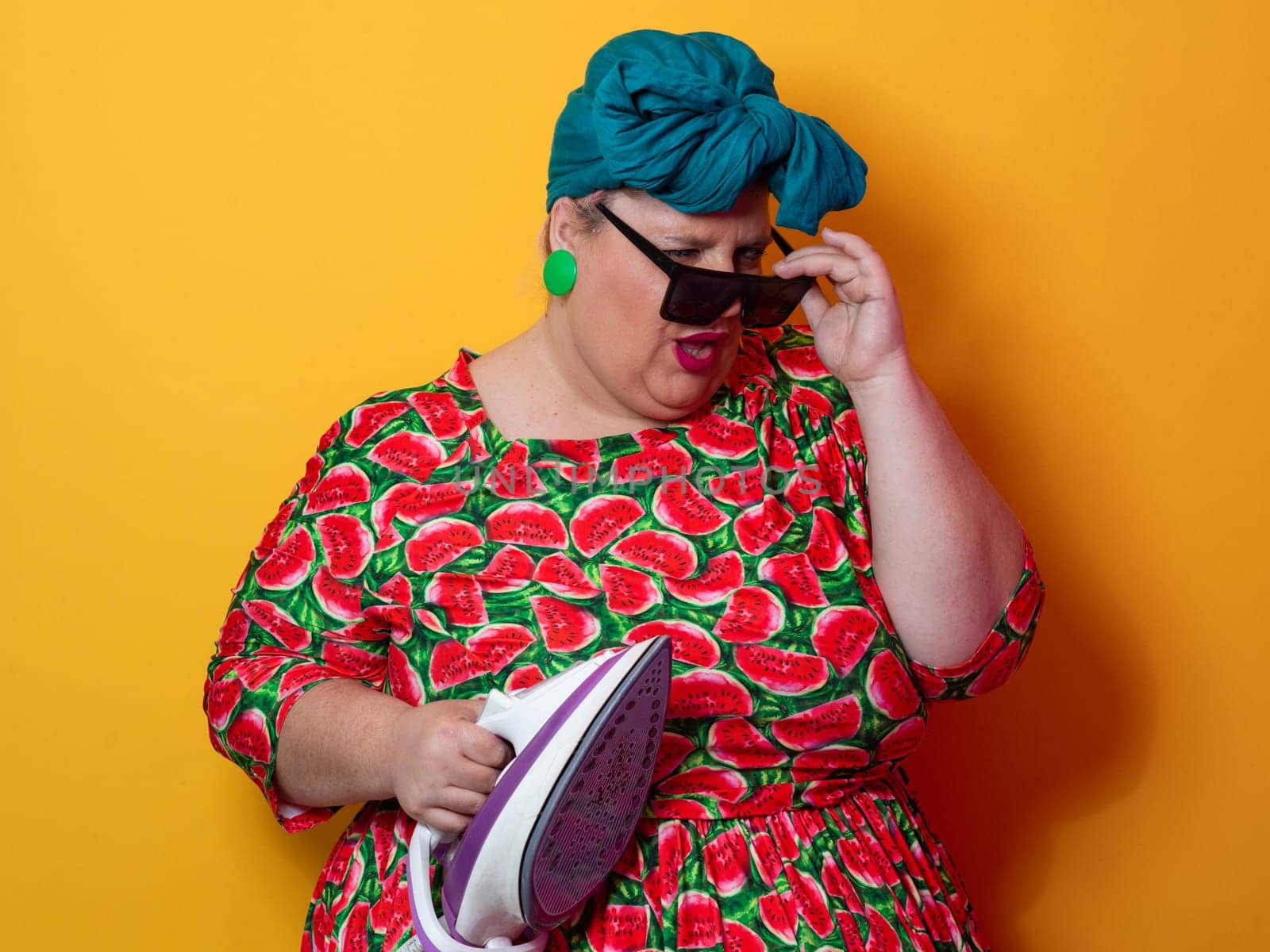 Plus size woman in a vibrant watermelon-patterned dress and teal headwrap, holding an electric iron and shouting with a humorous expression against a bright orange background. Conceptual and colorful portrait representing loudness, domestic chores, and quirky energy.