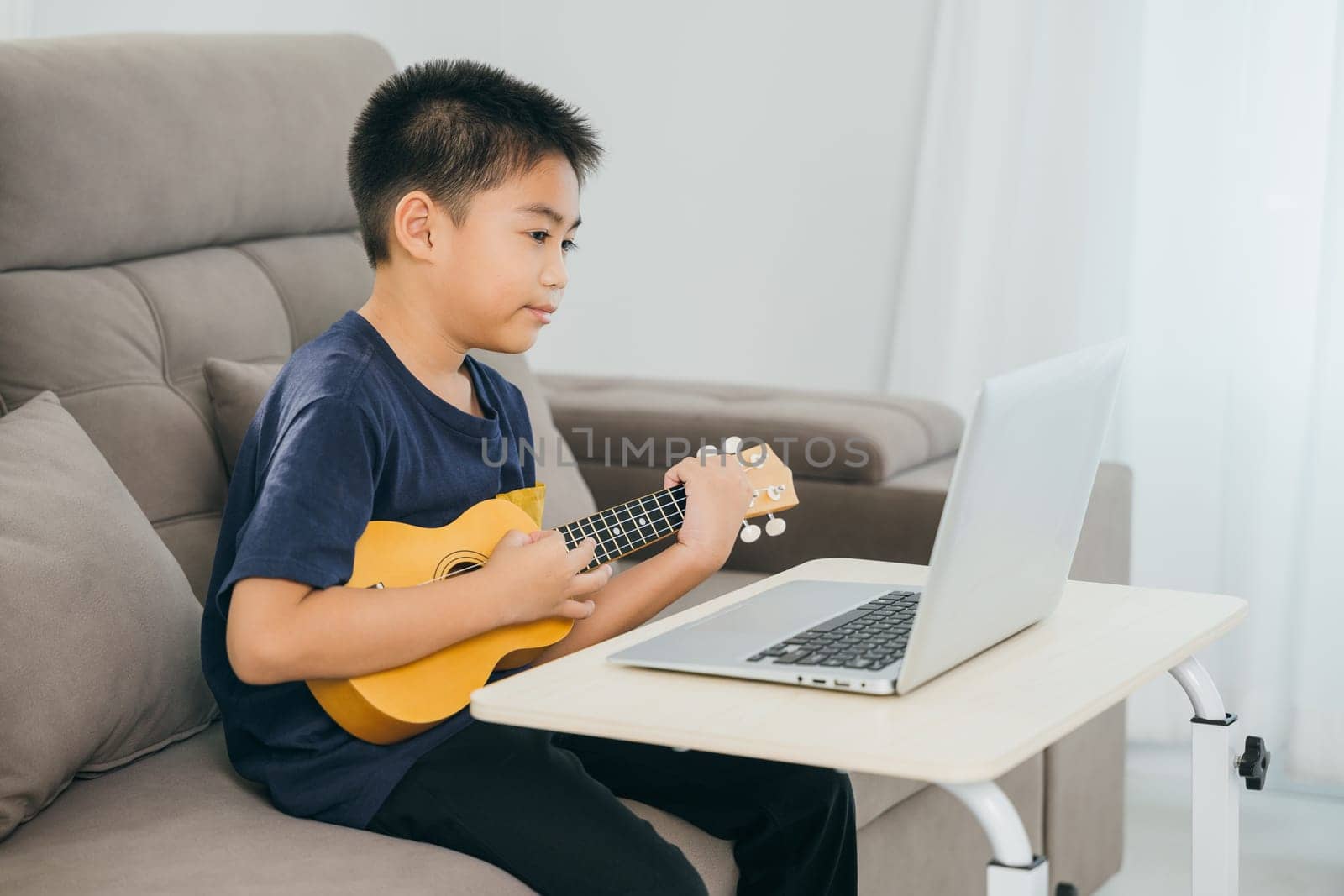 Boy learning ukulele on a sofa while watching a tutorial on a laptop. The cozy living room scene highlights the integration of technology and creative education in a modern home environment.