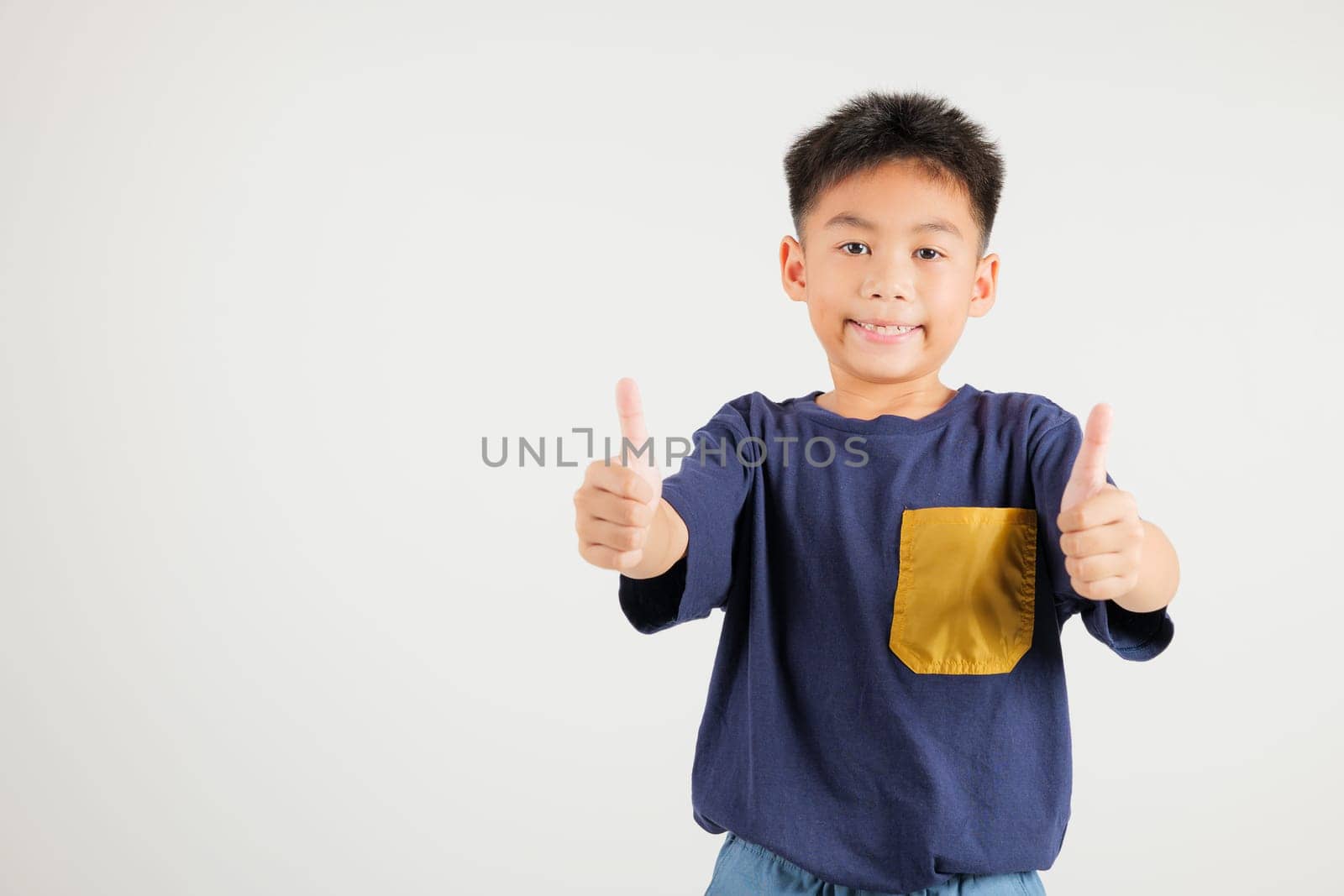 Confident Asian young boy in a studio portrait isolated on white background makes a thumbs up OK sign, symbolizing agreement and success. primary child smile radiates positivity and satisfaction
