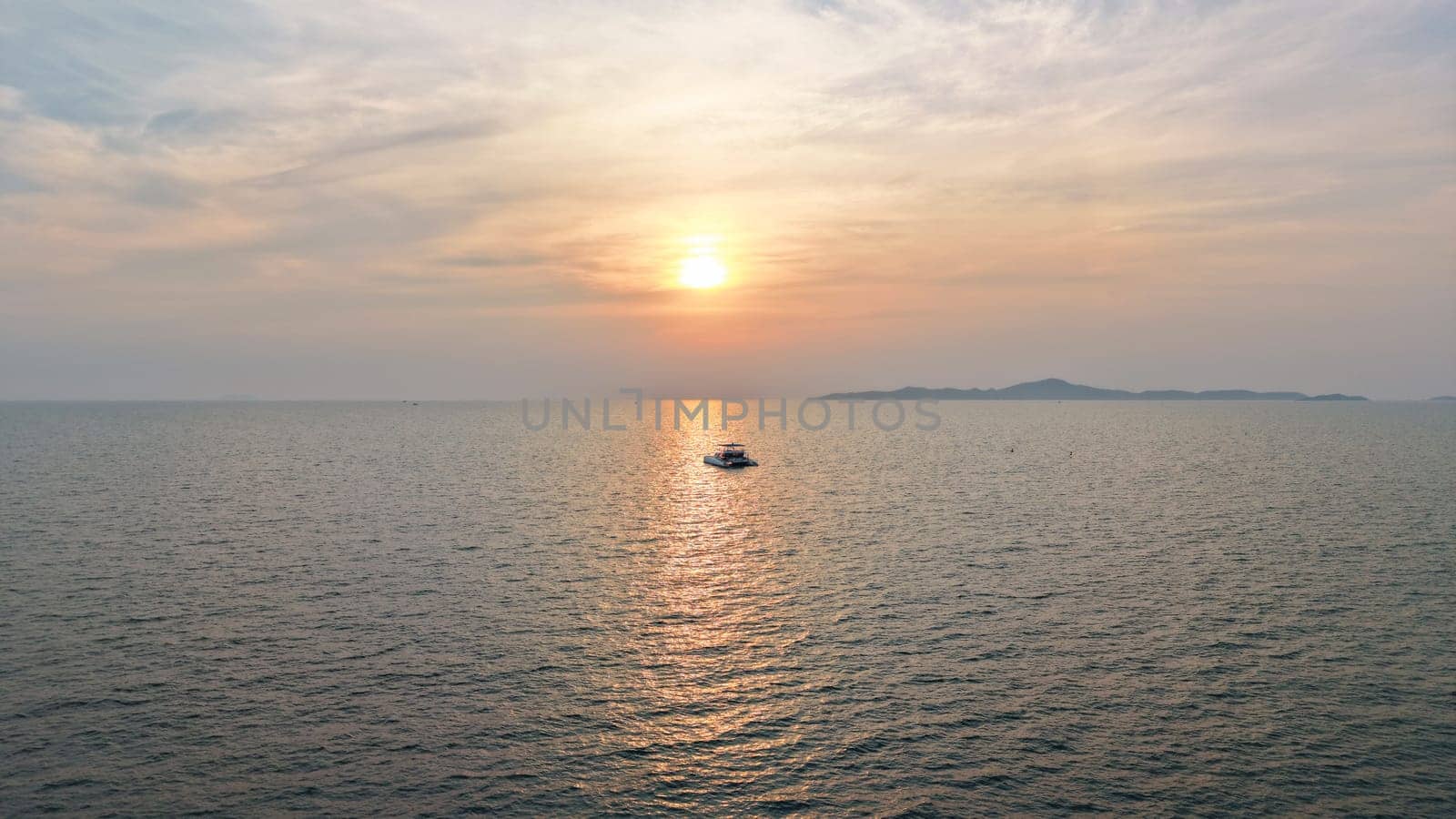 Golden sun setting over a tranquil ocean, casting a warm glow on the water as a catamaran sails peacefully towards the horizon, with a distant island adding to the serene seascape