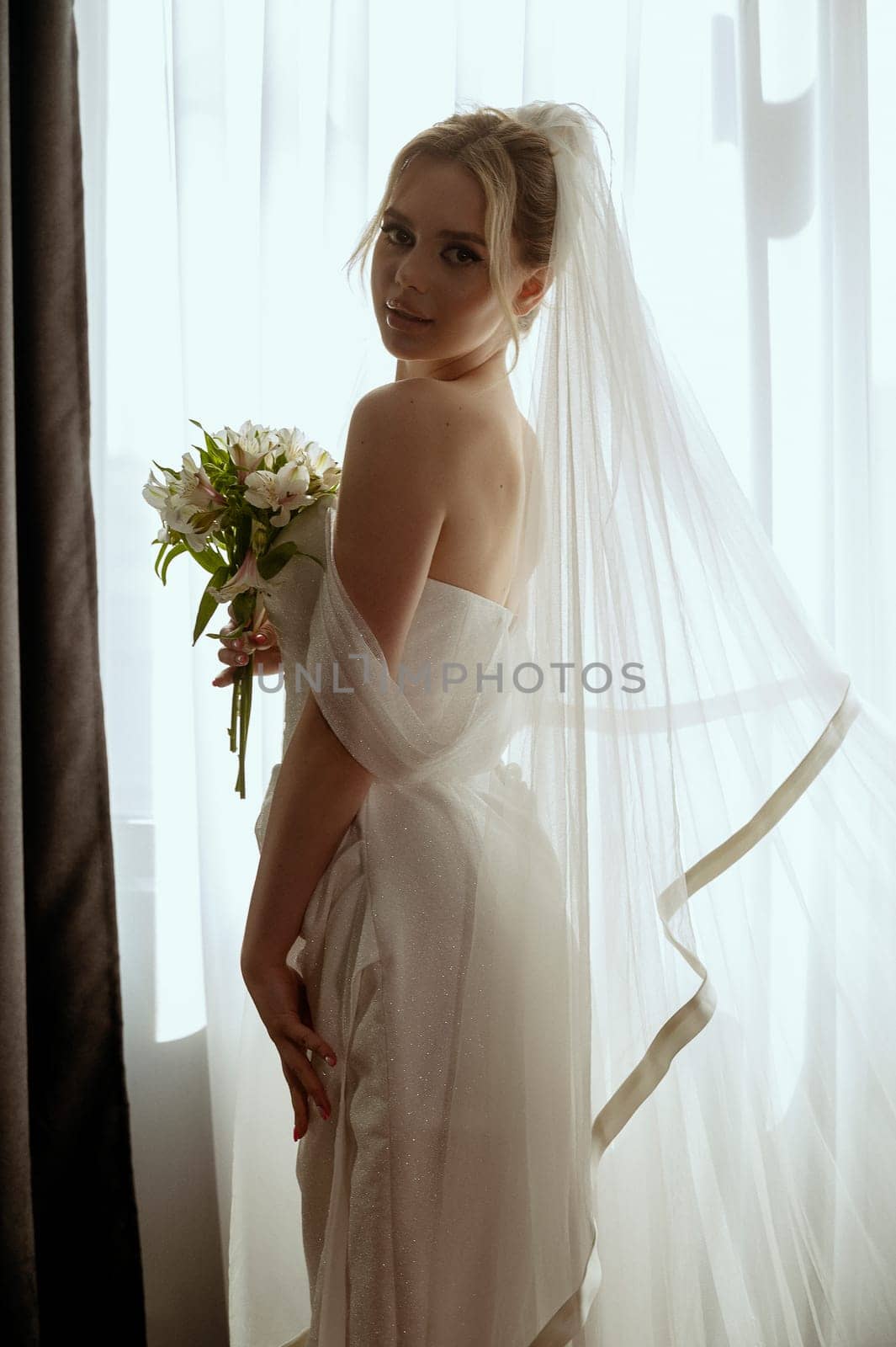 blonde bride in a long veil stands in a long white dress near the window with a bouquet of lilies in her hands. Minimalistic interior, hotel or house. bride in a long dress with a slit on the side