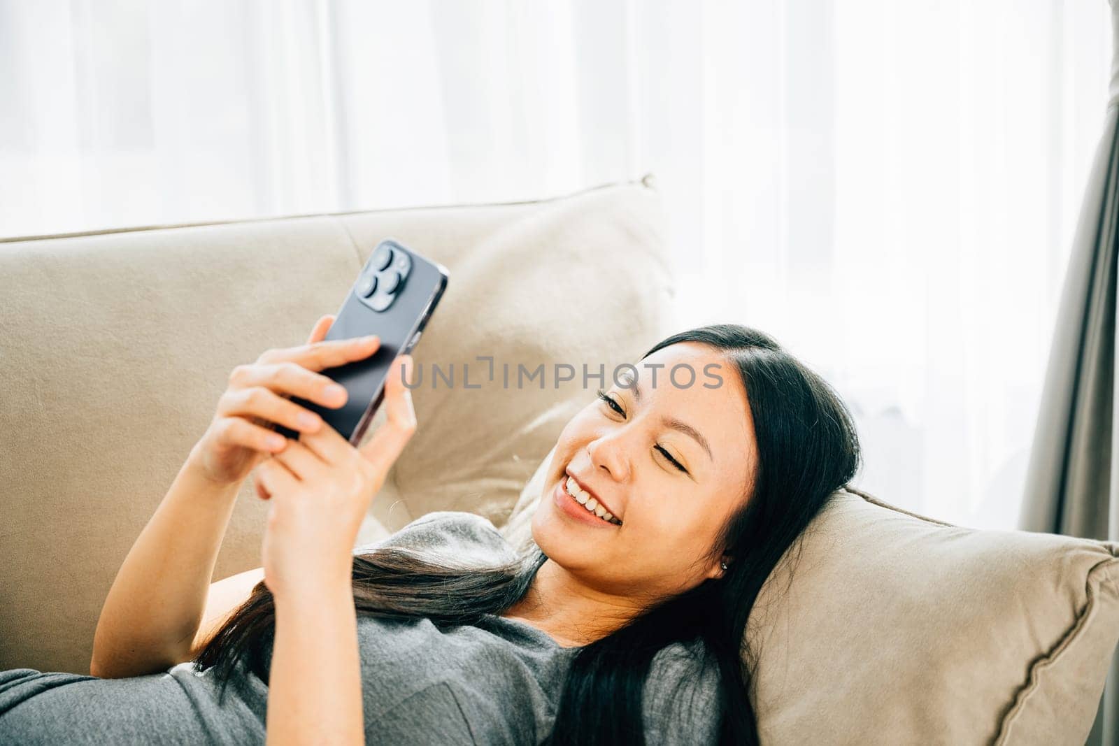 Young Asian woman relaxes on sofa texting on smartphone. Enjoying relaxation chatting and online shopping. Modern technology for communication and connection.