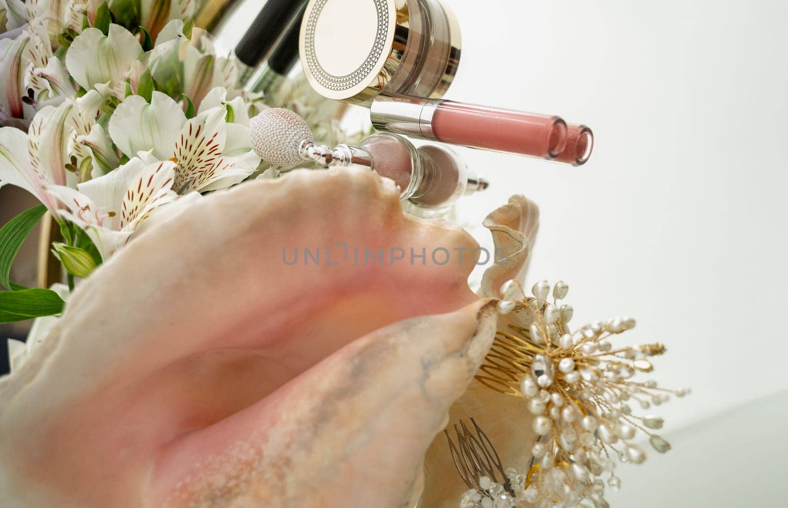 Pink lip gloss, highlighter, powder, flowers, pearl hair ornaments, makeup brush, large seashell on mirror background. Flat lay with space for text.