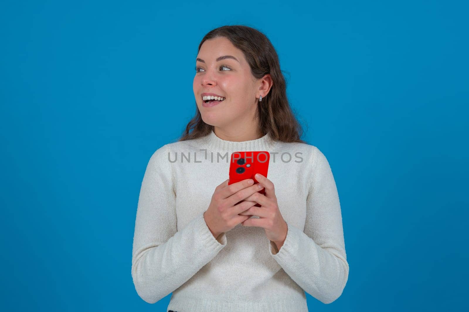 Happy young woman holding a red smartphone while looking to the side with a surprised expression. She stands in front of a bright blue background, expressing excitement and curiosity.