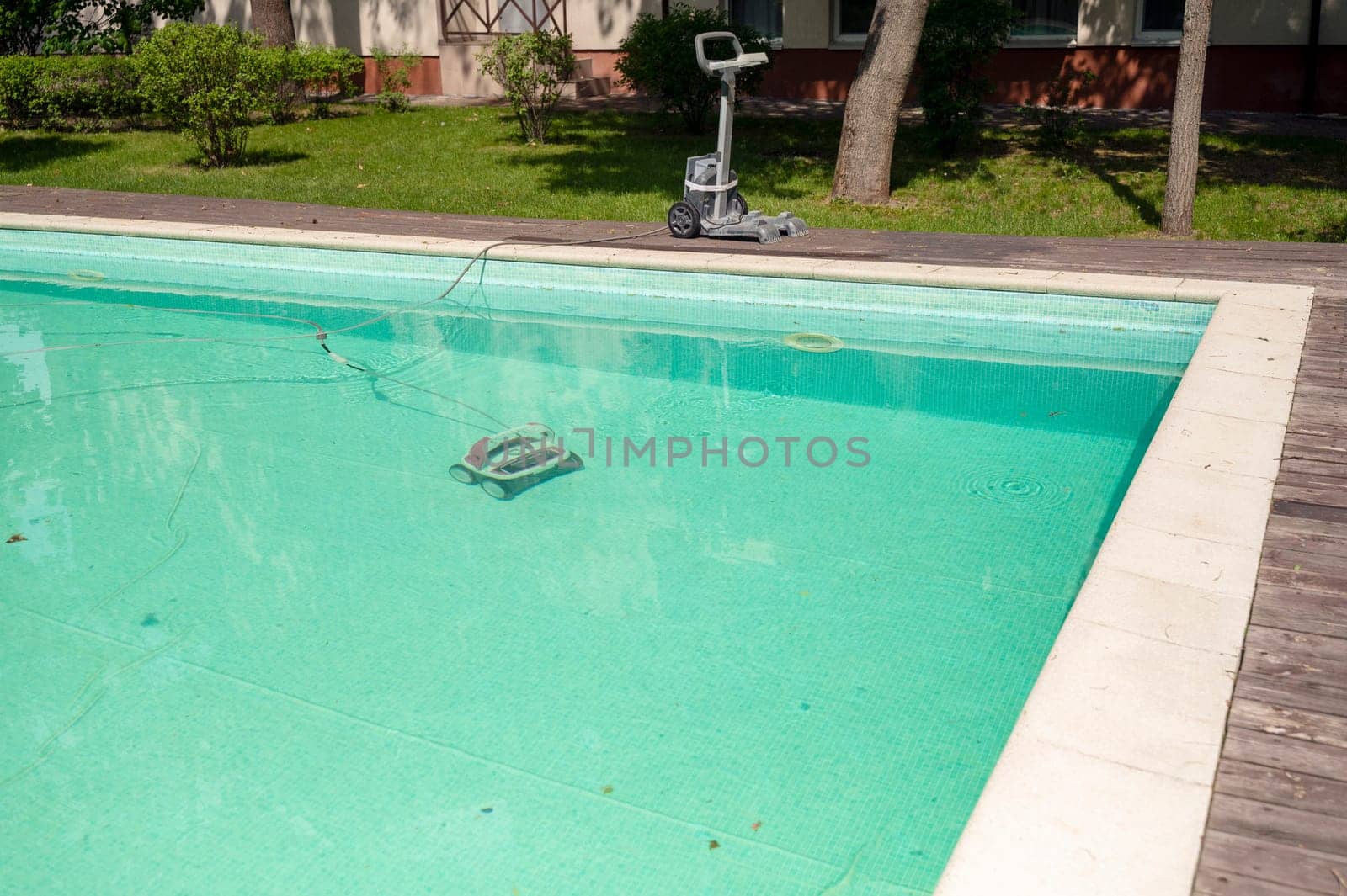 A swimming pool with a wooden deck around it, in a hotel garden, and water cleaning equipment. A pool cleaning machine is running in the pool. The equipment is in the water and nearby
