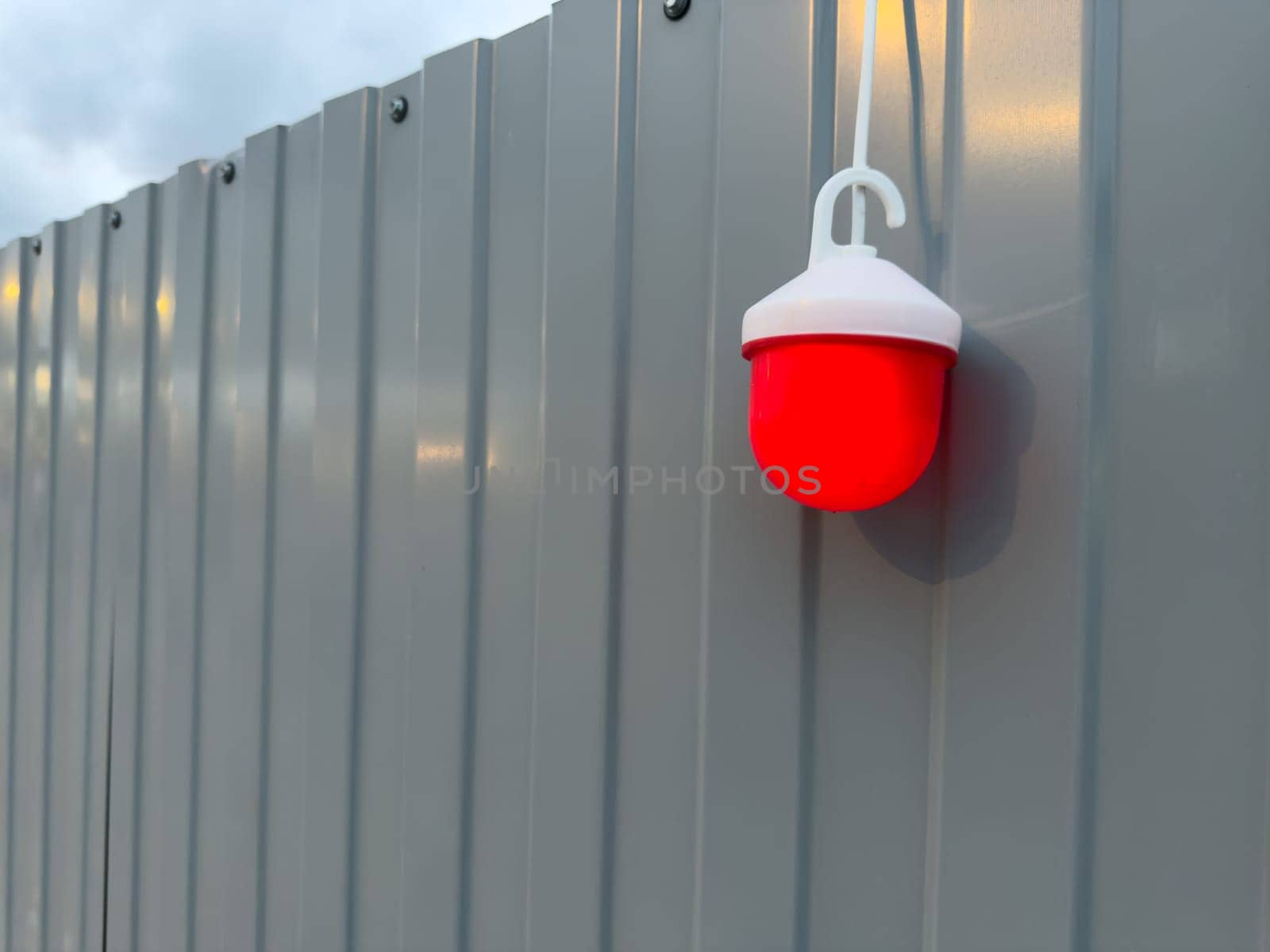 Close up of red warning light mounted on gray metal fence at construction site during twilight. The glowing lamp adds contrast and draws attention to safety and hazard awareness in public spaces