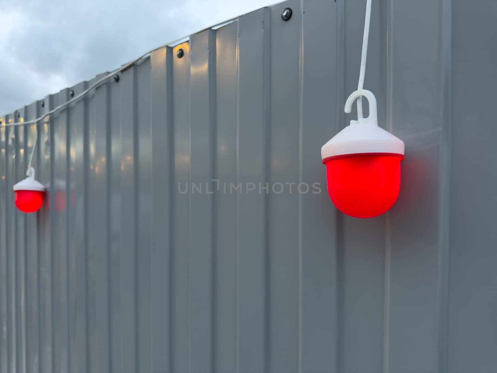Red warning safety light hanging on a gray metal construction fence at dusk, used for marking boundaries or hazards in urban development and renovation zones