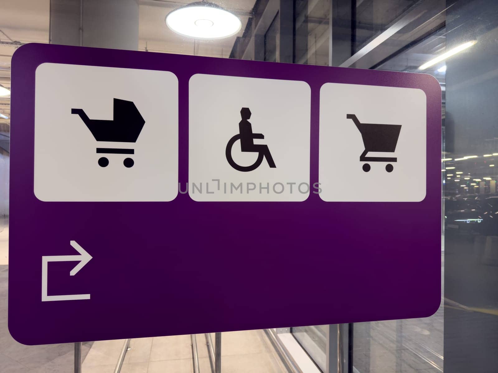 Purple directional sign with black icons indicating areas for baby strollers, wheelchair users and shopping carts, placed in modern parking facility with glass walls and lighting reflections
