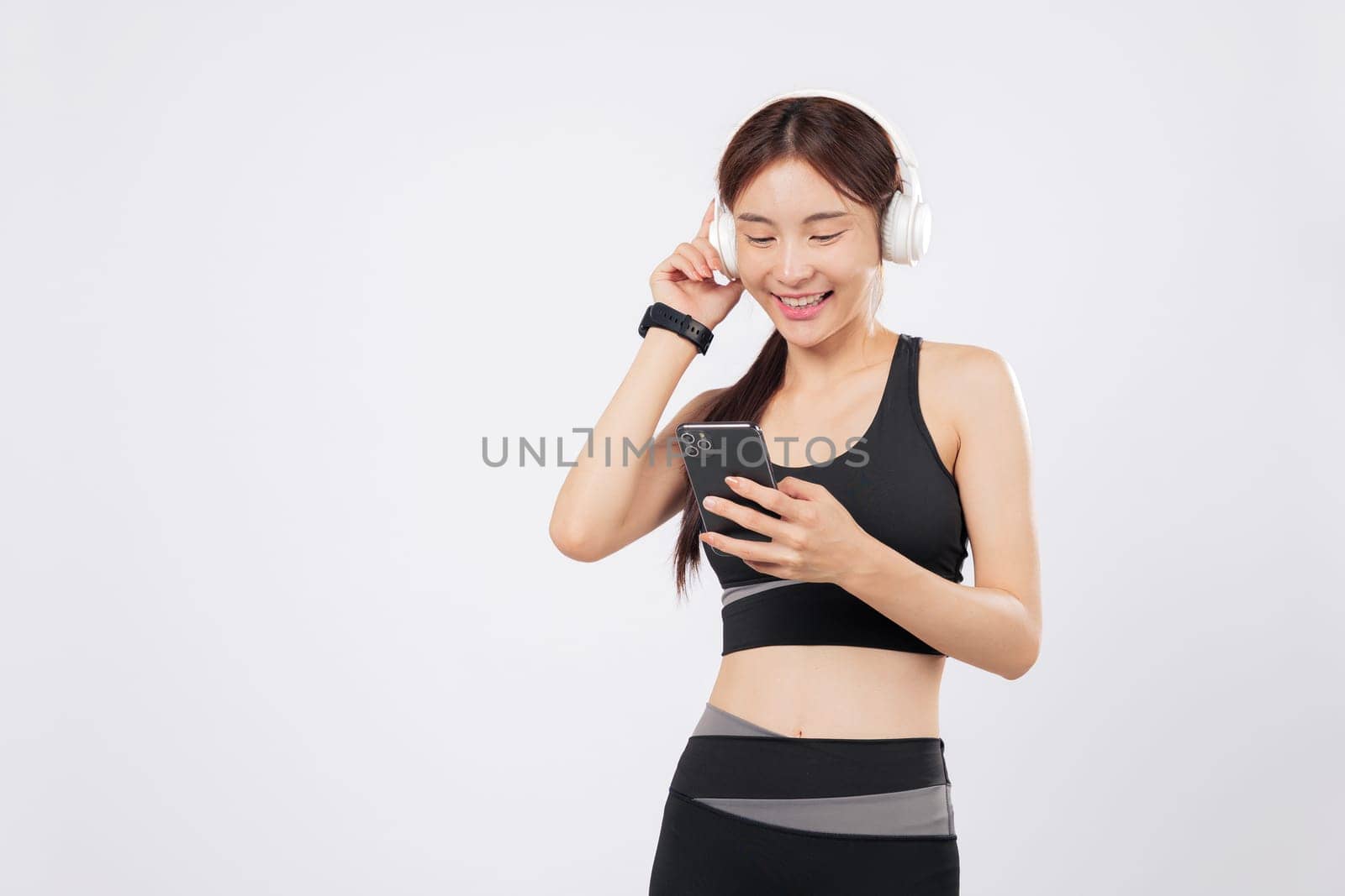Holding her phone while browsing application music on the phone, this athletic Asian woman in sportswear listens with joy through her headphones during a workout. isolated studio white background