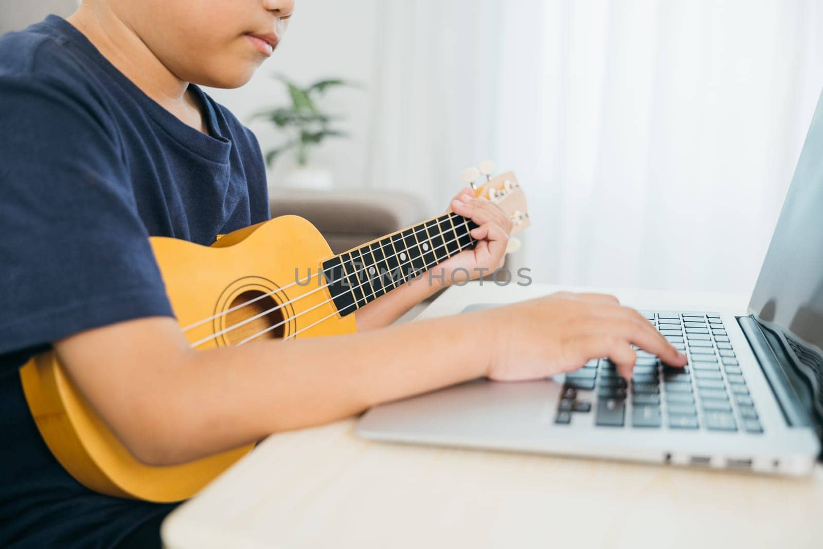 Hands of a kid boy playing a ukulele, with a laptop nearby showing an online music course. The living room scene creates a cozy and educational environment for his practice and creativity.
