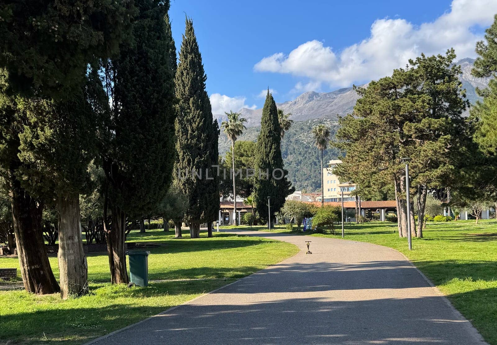 Budva,Montenegro-April 11,2025:A wide paved path winds through a green park with tall cypress and palm trees, and a backdrop of mountains. High quality photo