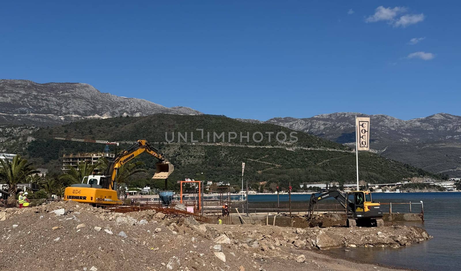 Budva,Montenegro-April 11,2025:A Volvo excavator working on a waterfront construction site with mountains and blue sea in the background. There is a flag with the word COCO next to it. High quality photo