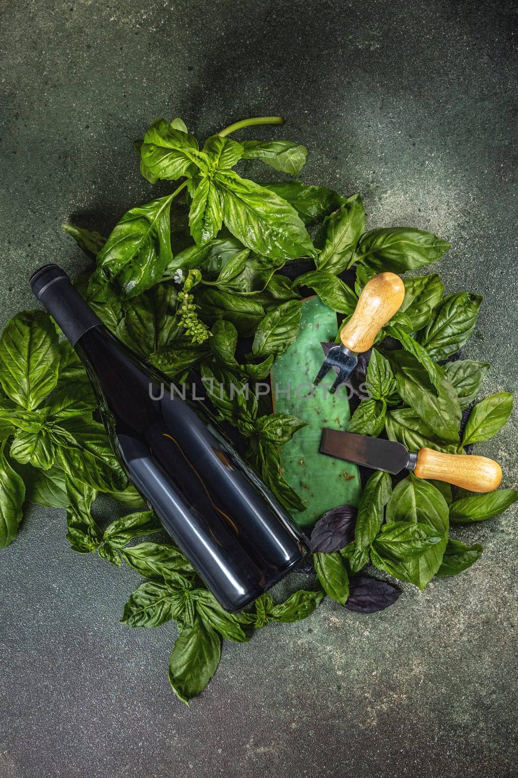 Wine bottle lying on fresh basil leaves near cheese tools and green cheese on dark background, celebrating flavors and aromas
