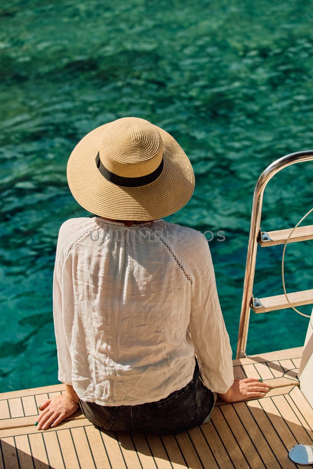 A girl in a hat sits on the edge of a sailing boat and enjoys the views and azure color of the sea on a sunny day. High quality photo
