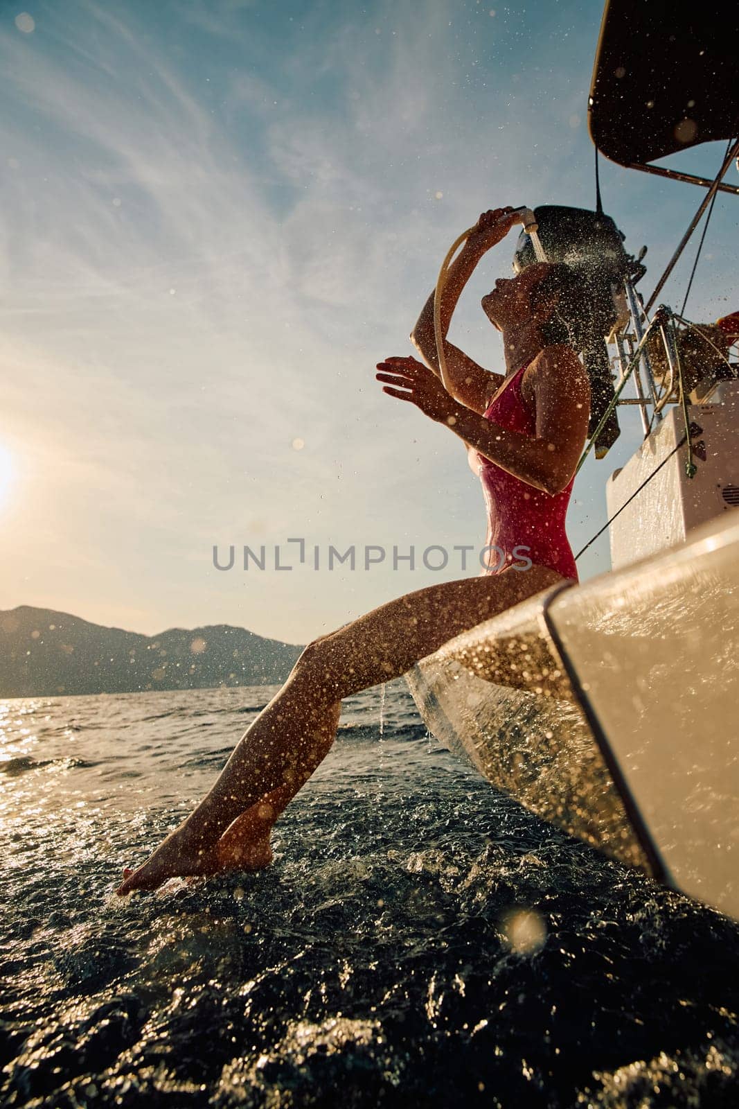 A girl in a swimsuit at the stern of a sailing boat takes a shower after swimming in the sea at sunset, scattering luminous splashes of water, the edge of a sailing boat, azure color of the sea. High quality photo