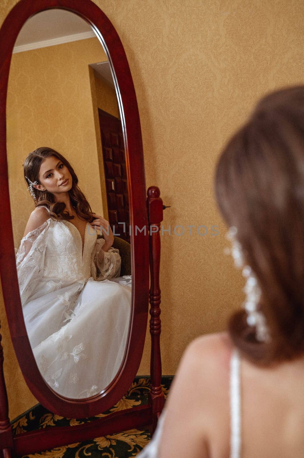 Bride in long white dress standing in front of mirror in hotel room before wedding. Her long hair is decorated with white flowers. Back view, White corset with lacing. Foreground is blurred.