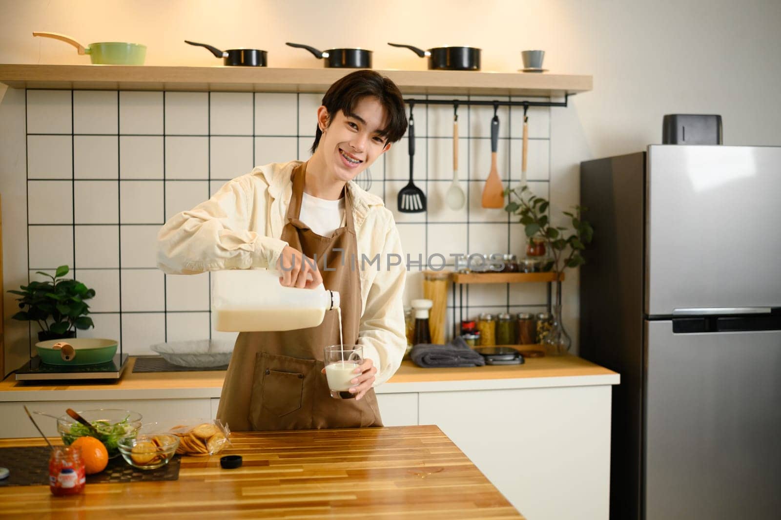 Cheerful young man pours fresh milk into a glass, standing in a modern kitchen.