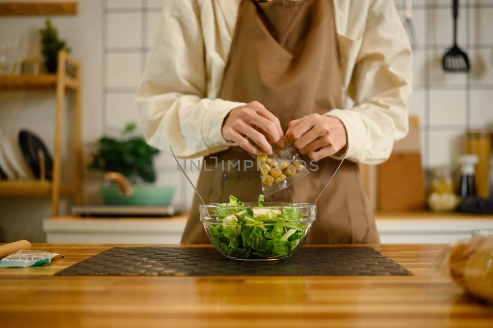 Close up of a man in an apron adding croutons to a bowl of fresh green salad.