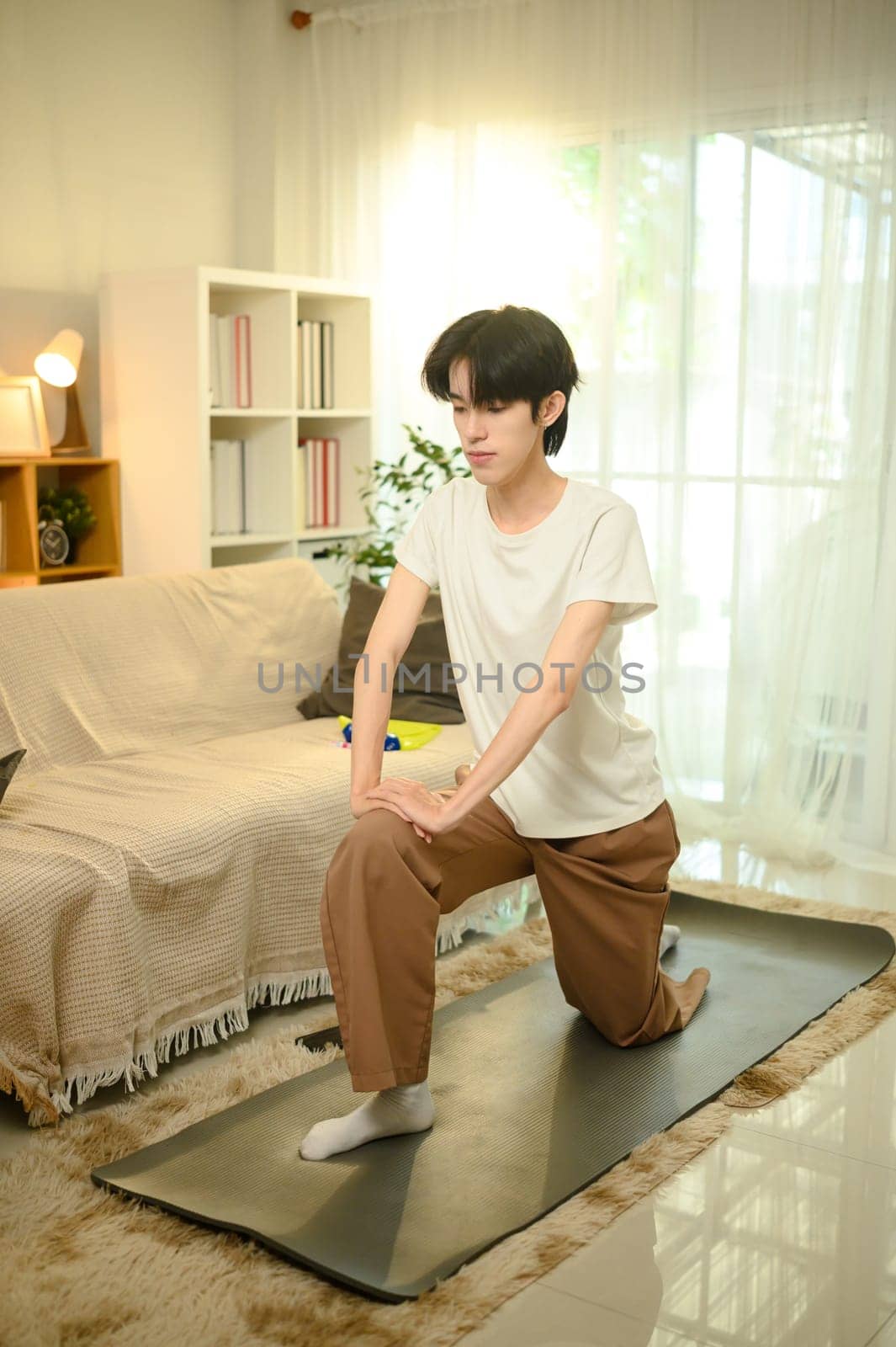 Smiling young asian man practices yoga on a mat in his living room.