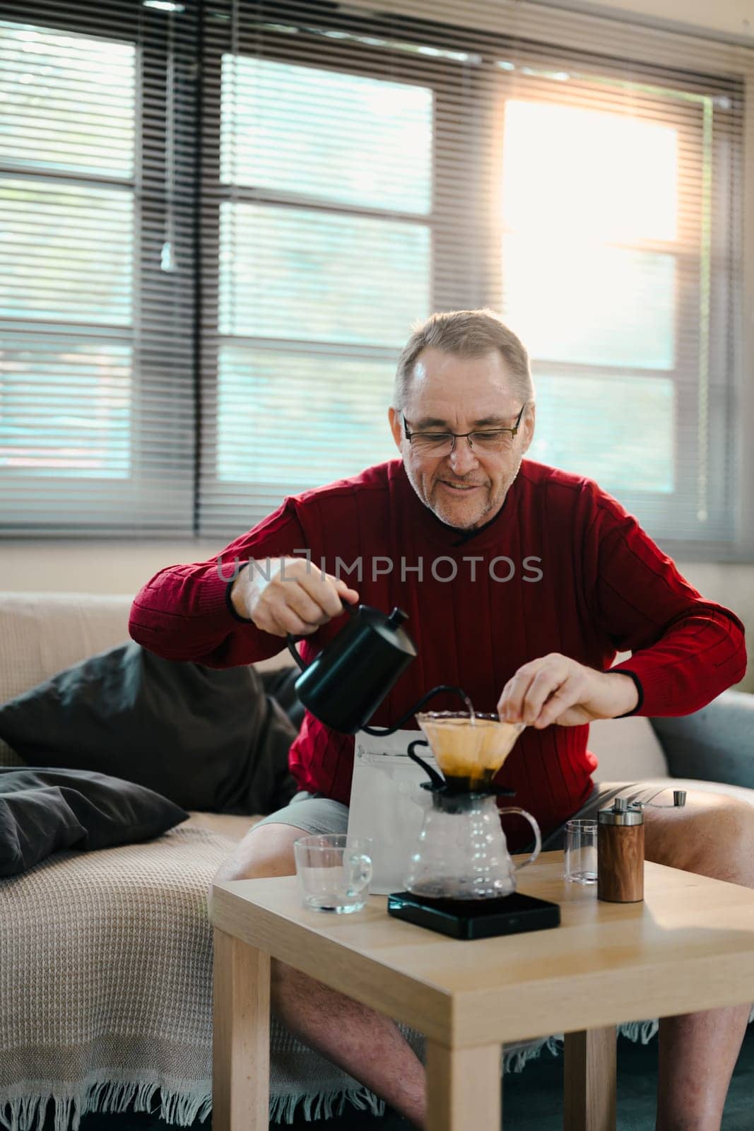 Happy elderly man carefully making pour-over coffee at home with a glass dripper and kettle.