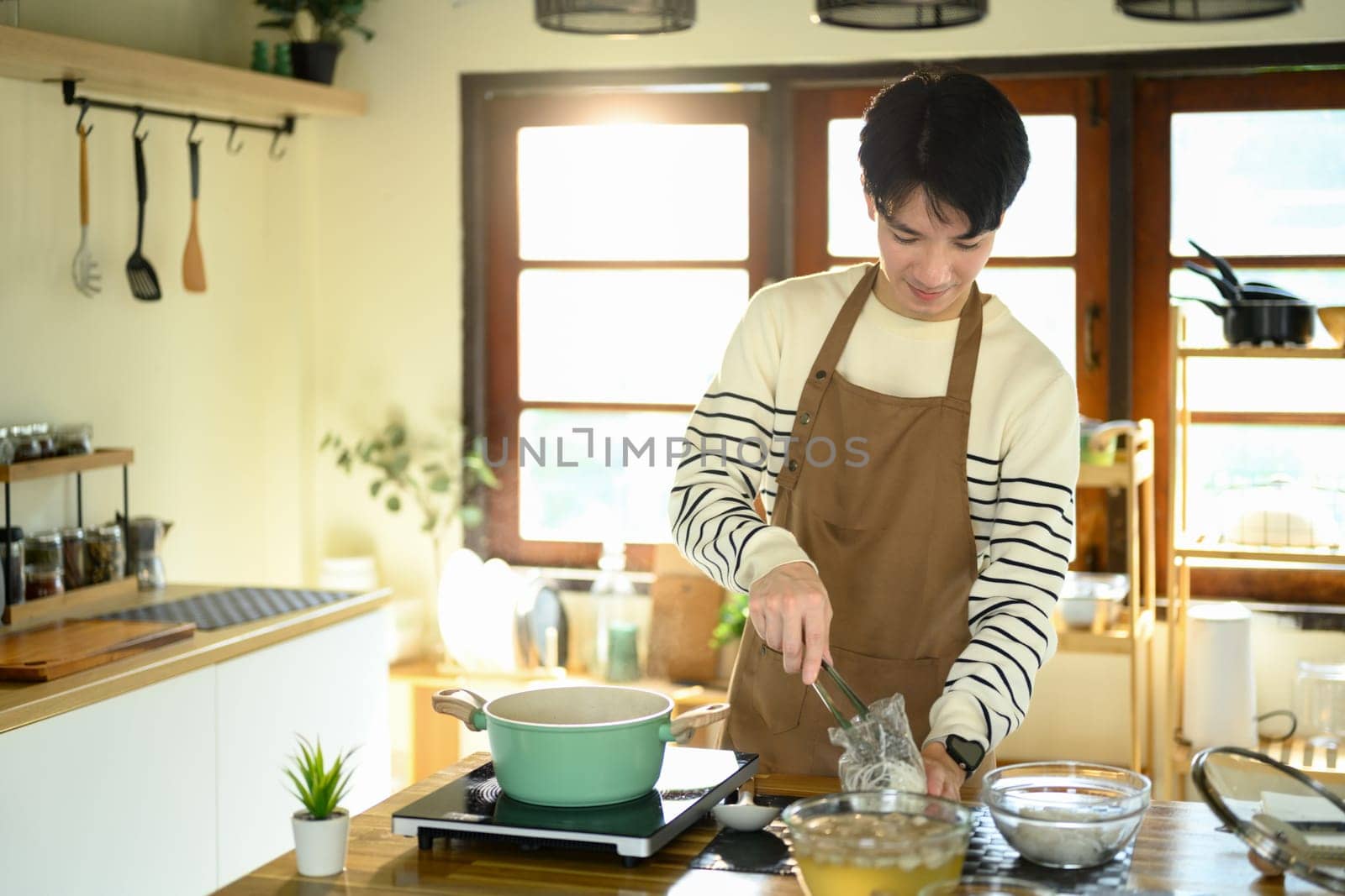 Smiling man in an apron prepares noodles over a stove in warm modern kitchen.