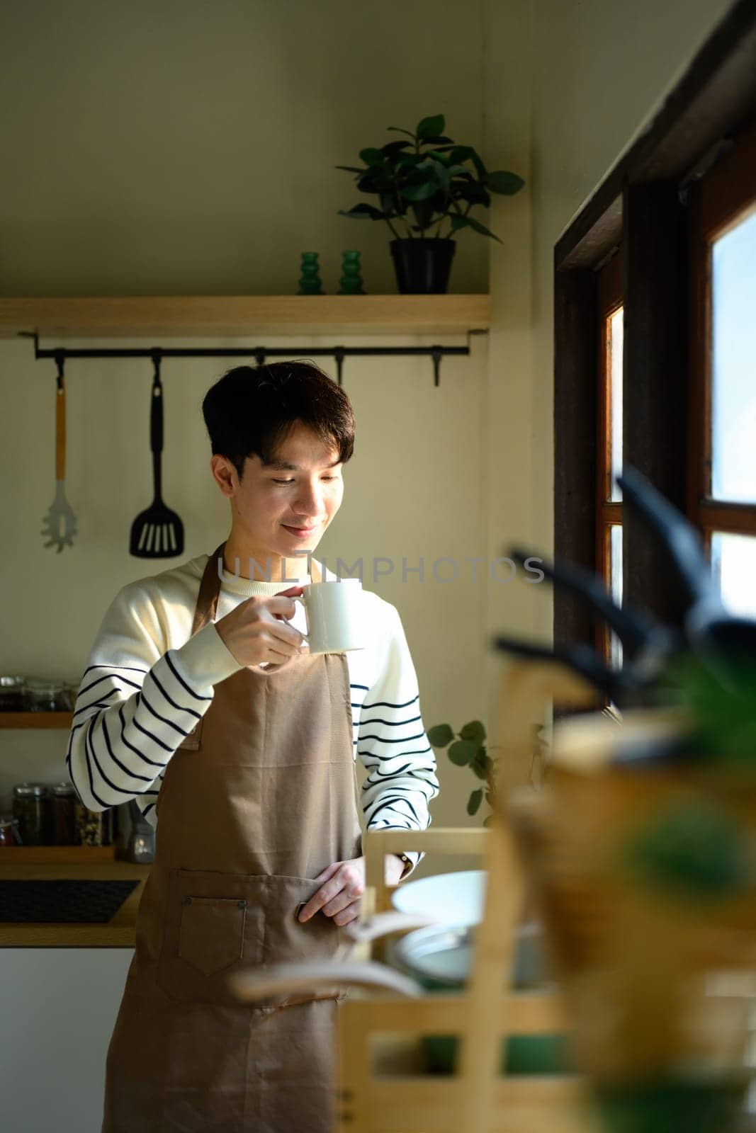 Man in an apron stands by the window, savoring a warm drink in a sunlit kitchen.