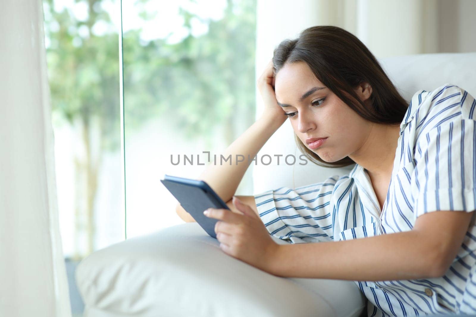 Bored and disappointed woman reading ebook sitting on a couch at home